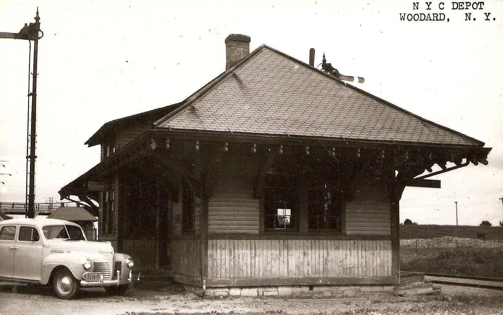 Old black and white photo of a train depot building with a vintage car parked in front. The signage reads "N Y C DEPOT WOODARD, N. Y."