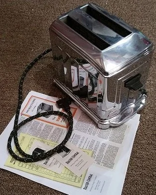 A chrome bread machine with a black power cord, sitting on papers and manuals on a brown carpet.