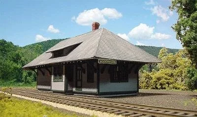 Vintage train station building with a pitched roof, situated next to railroad tracks in a lush green area.