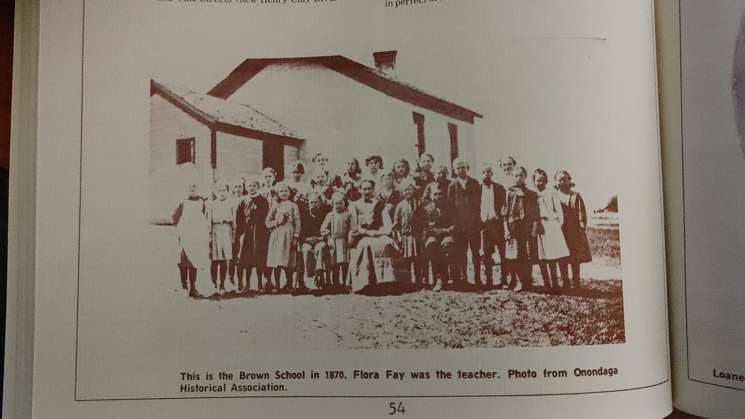 A sepia-toned photograph of a large group of children and a female teacher standing outside a wooden schoolhouse in 1870. The children are dressed in period clothing, with some holding books and others standing with their hands at their sides. The sc