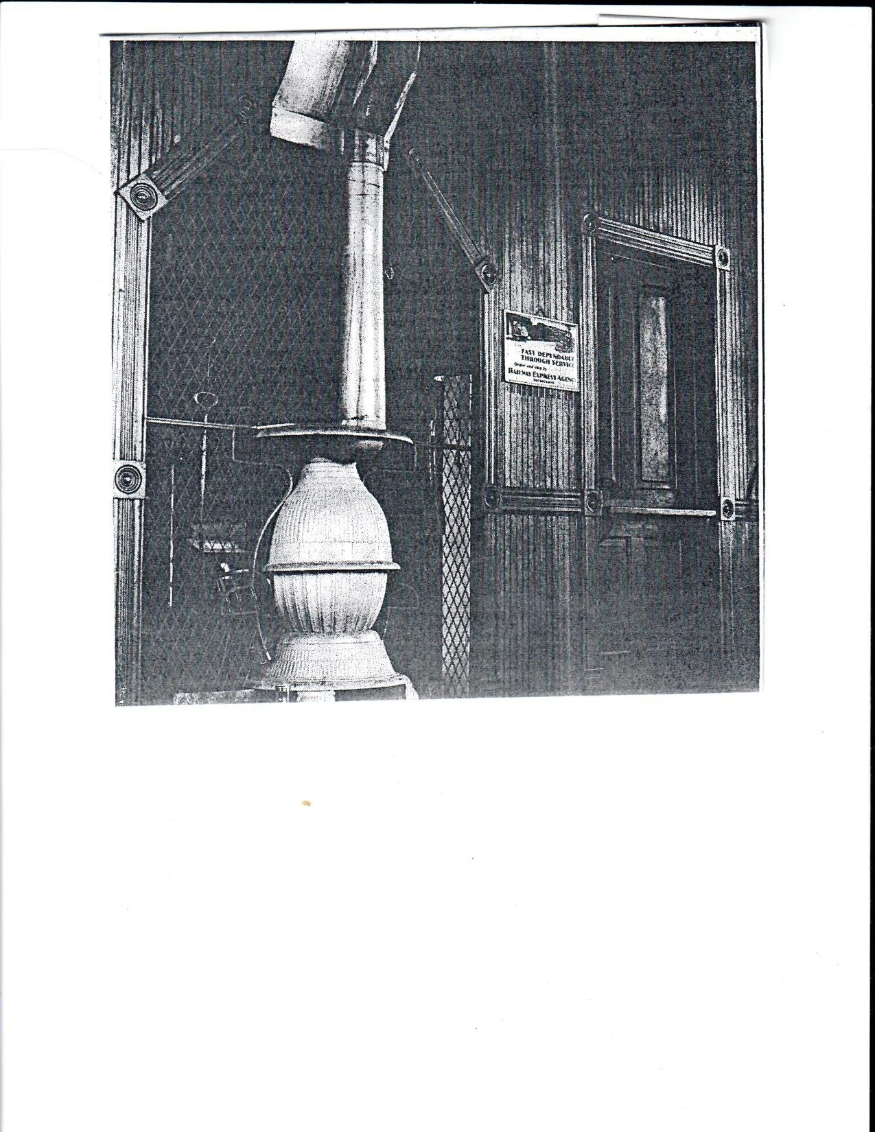 Black and white photo of a stove with a tall chimney pipe, in front of a wooden wall with a door and a sign that reads 'Fast Delivery Through Service' and other unreadable text.