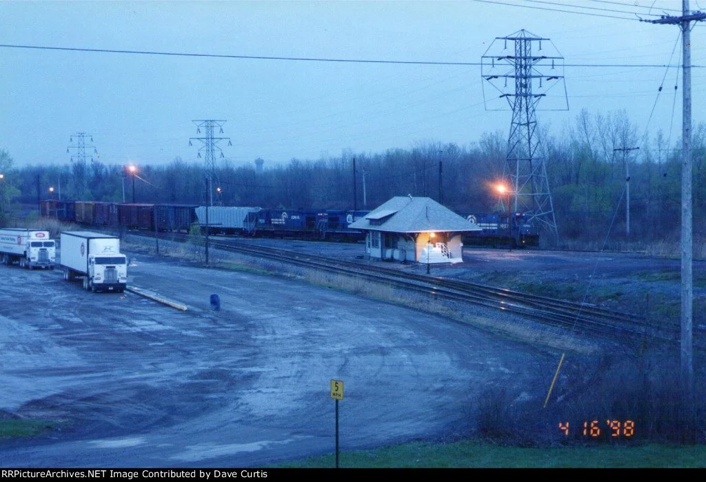 A train passing by a small train station with trucks parked nearby in a mostly empty parking lot during dusk.