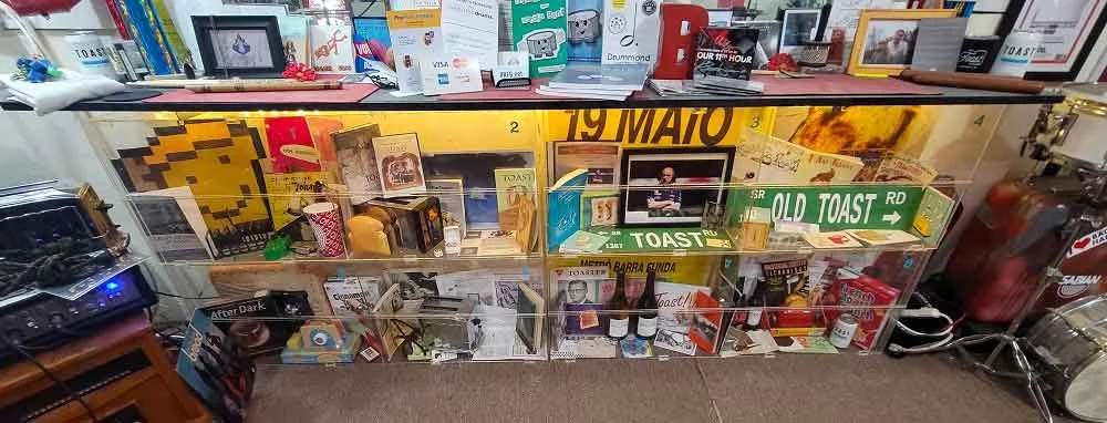 Display case with various memorabilia, photographs, and signs, including a road sign that reads 'OLD TOAST RD' and a yellow sign with the month 'MAYO'.
