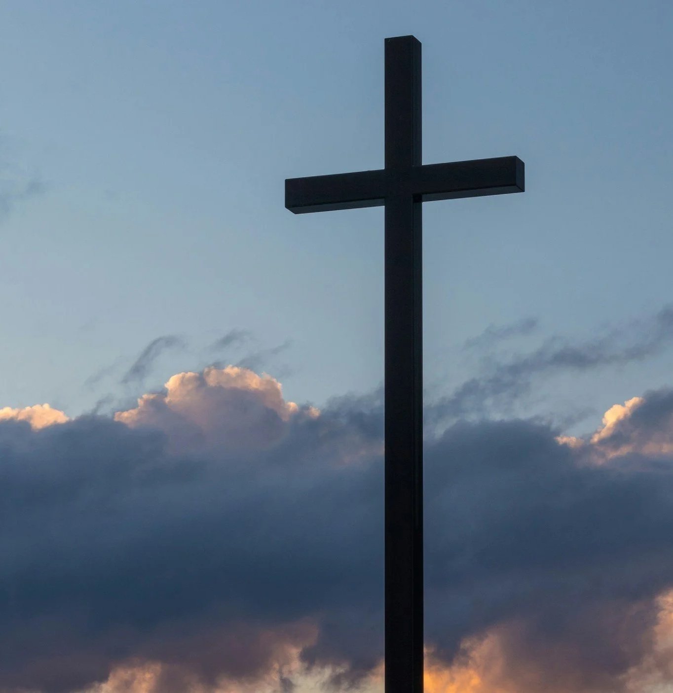A dark cross silhouette stands against a cloudy sky with hints of sunset colors.