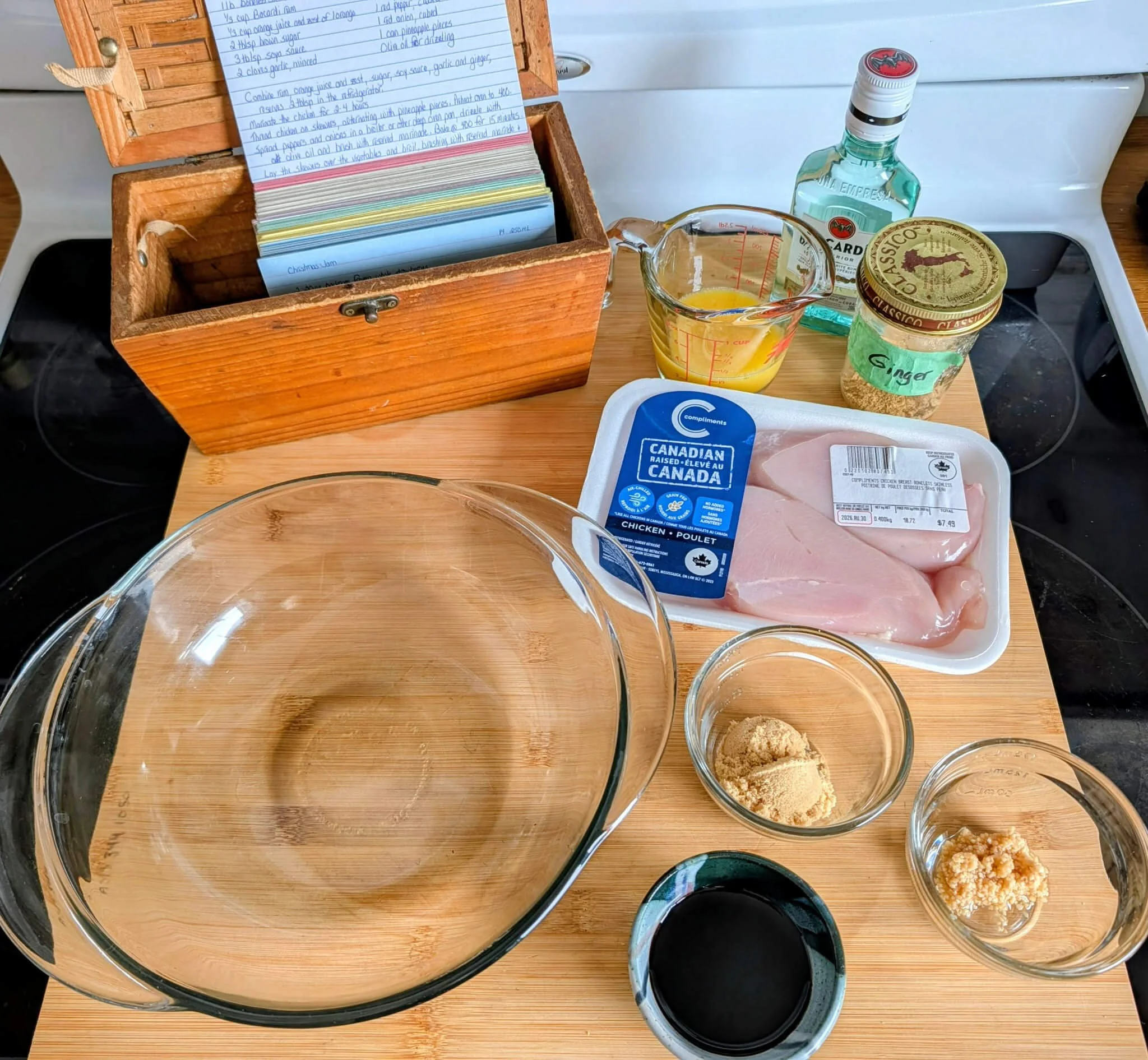 Ingredients for Caribbean chicken on a wood counter — raw chicken breast, orange juice, brown sugar, soy sauce, minced garlic, and ground ginger, with Sue's recipe box in the background