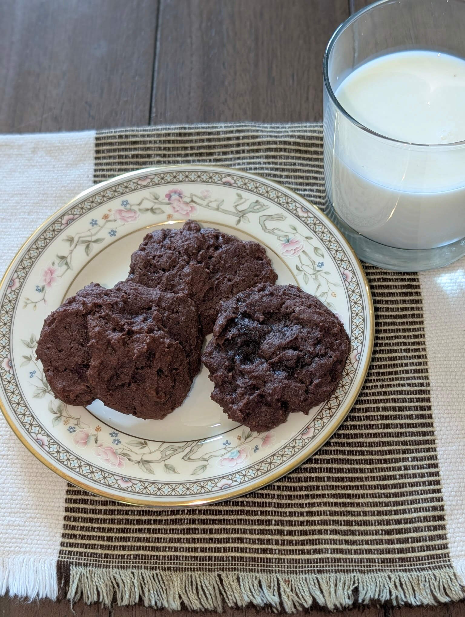 Four baked double chocolate chip cookies on a floral china plate with a glass of milk