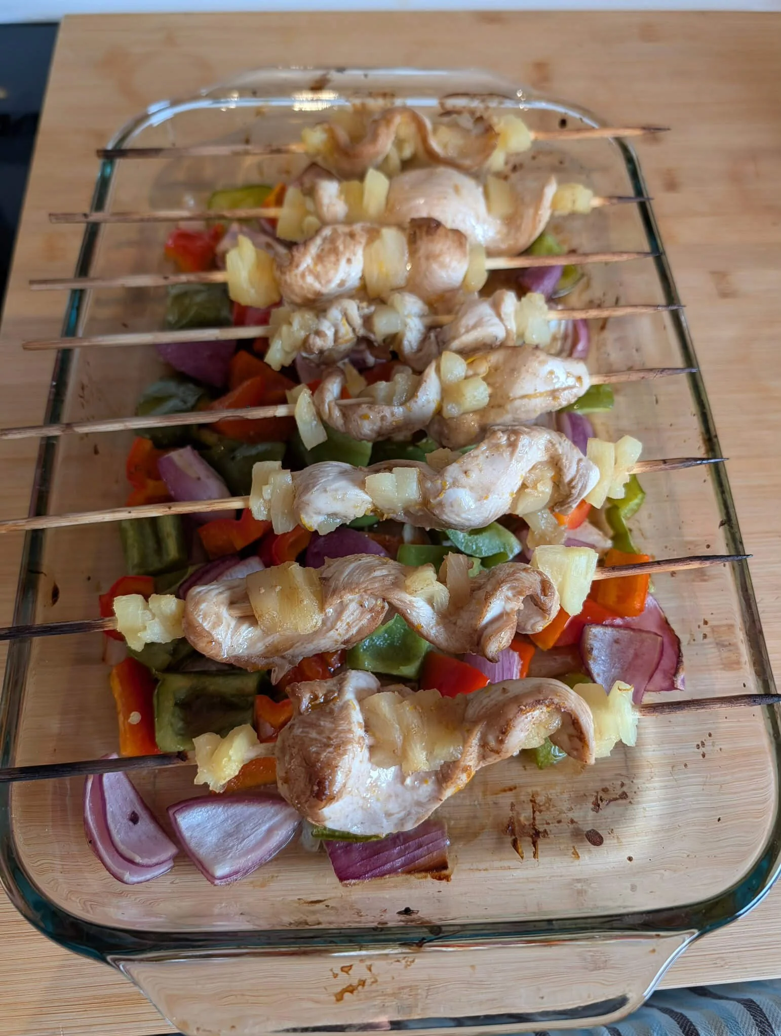 Baked rum-marinated chicken and pineapple skewers with caramelized edges, resting on a rack over roasted peppers and onions in a glass dish