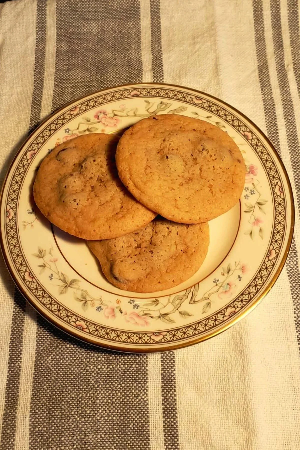 Three homemade chocolate chip cookies on a floral china plate on a striped linen cloth