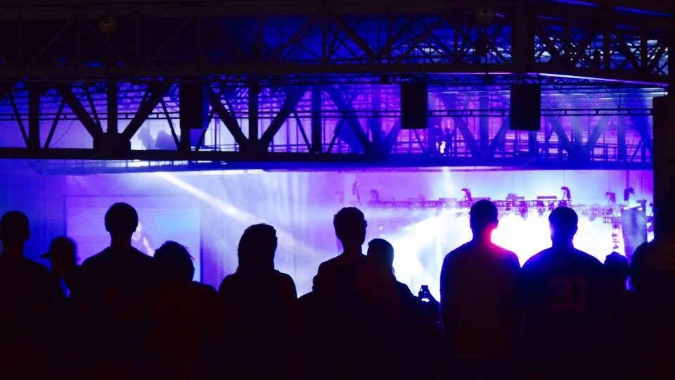 A foreground Silhouetted image of patrons at a music concert venue (The Pavilion in Charlotte), mid ground - purple and white lights shine brightly on stage in the back of the image
