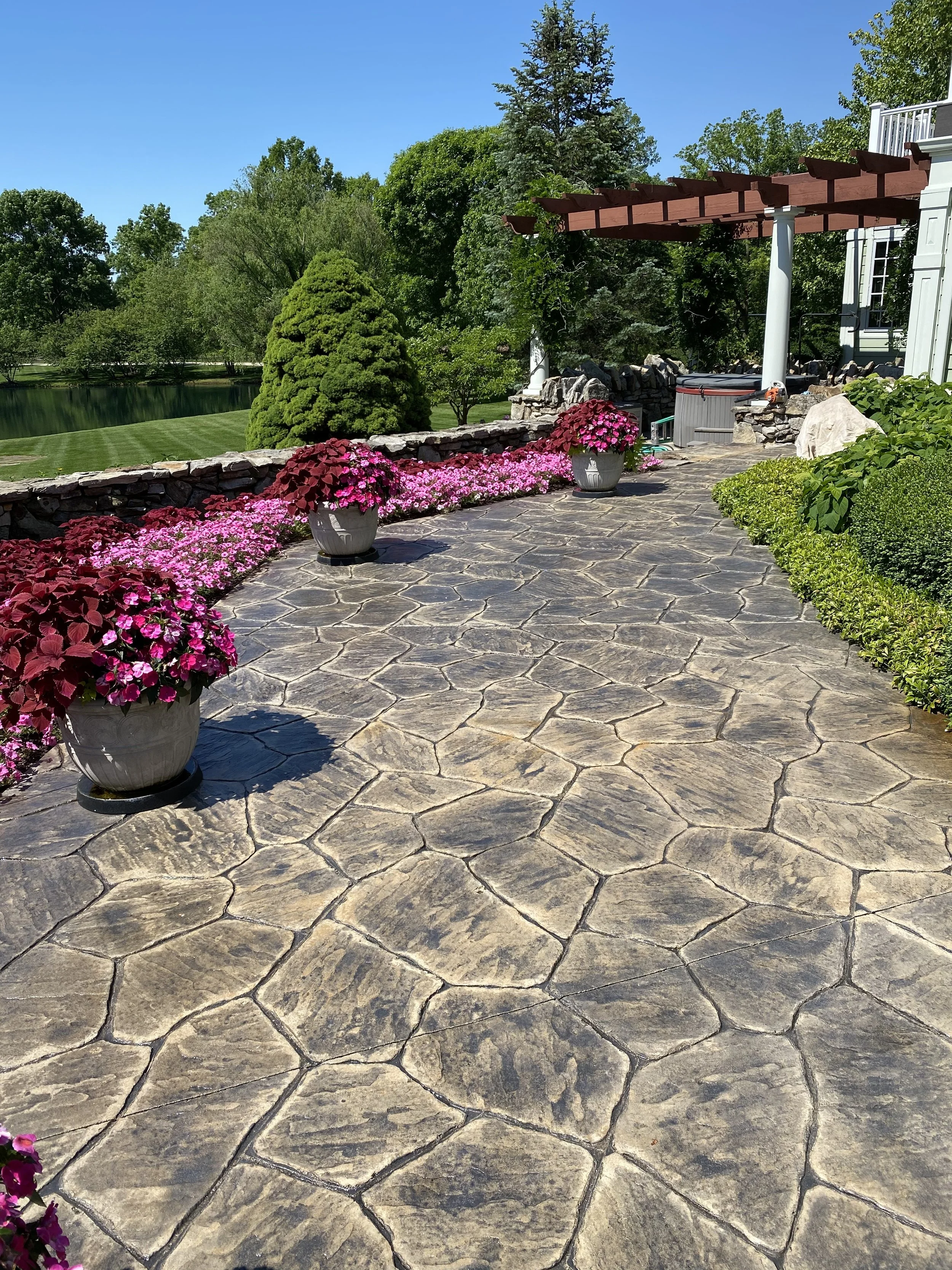 Pandus stone patio with potted pink and purple flowers, green shrubbery, trees, and a clear blue sky in a garden setting.
