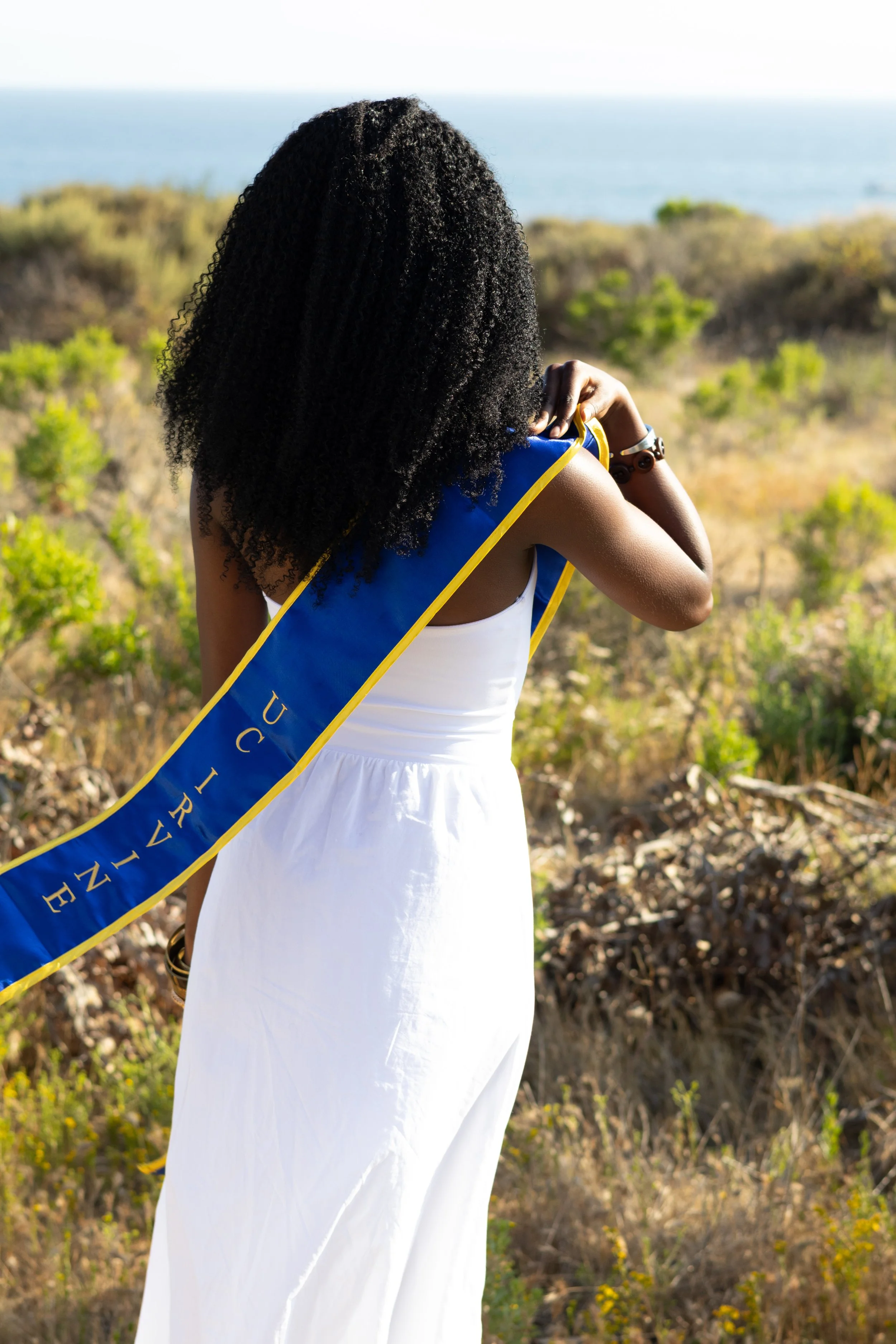 Woman with curly hair wearing a white dress and a blue sash that says 'UCRIVE' standing outdoors near shrubs with the ocean in the background.