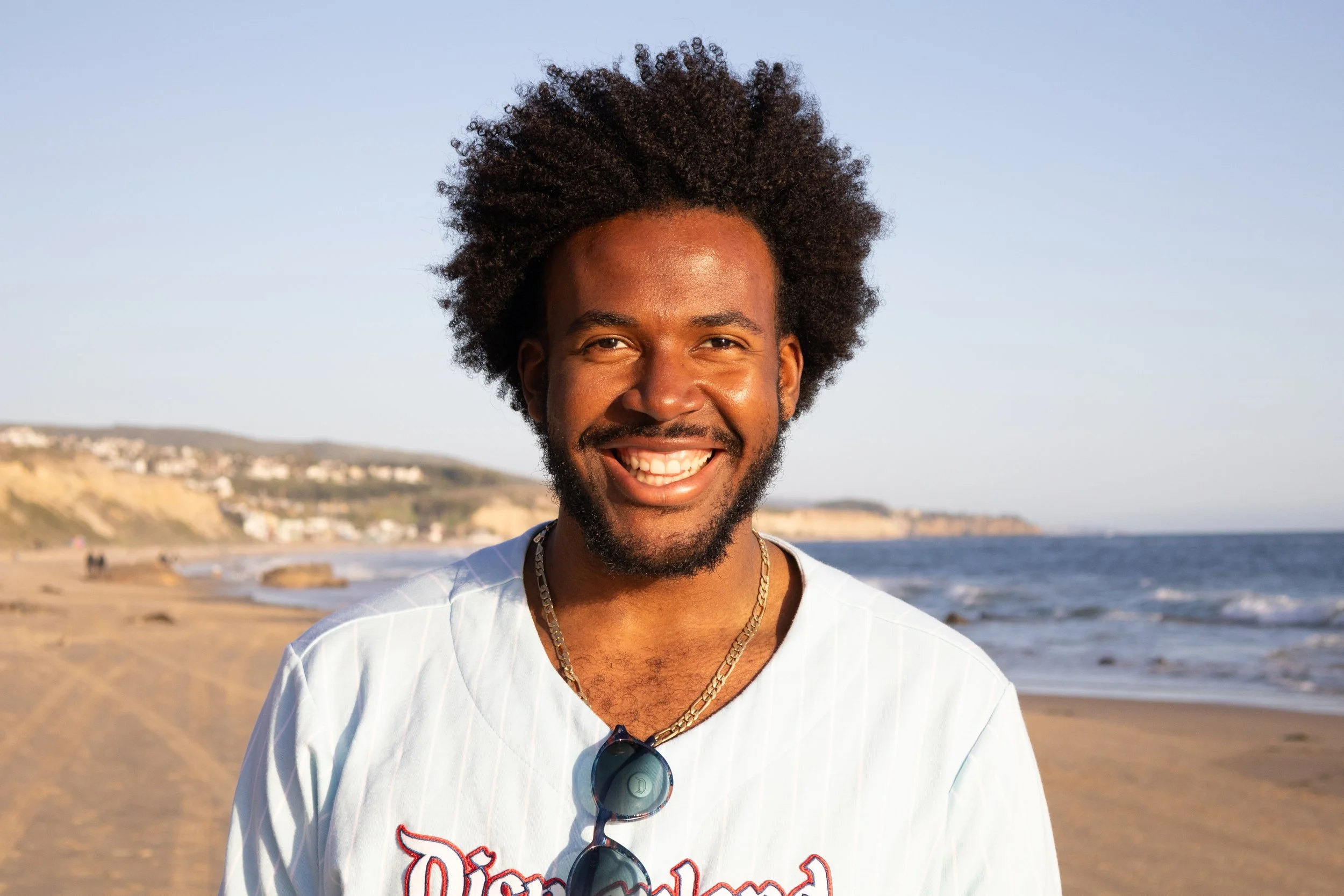 Smiling man with afro hairstyle and beard standing on a beach with ocean and cliffs in the background.