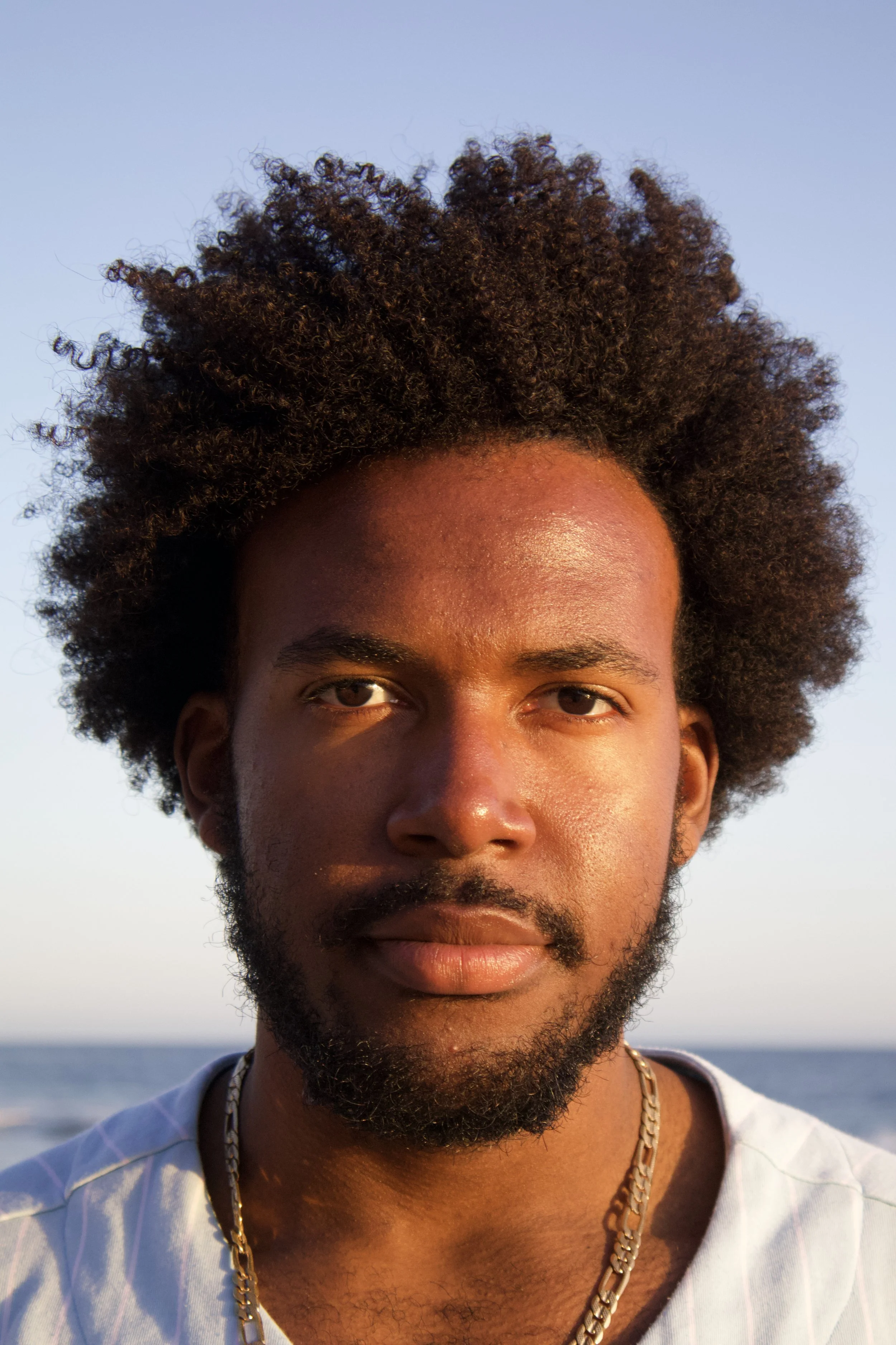 A close-up portrait of a man with facial hair, curly hair, and wearing a gold chain, standing outdoors during sunset near the ocean.