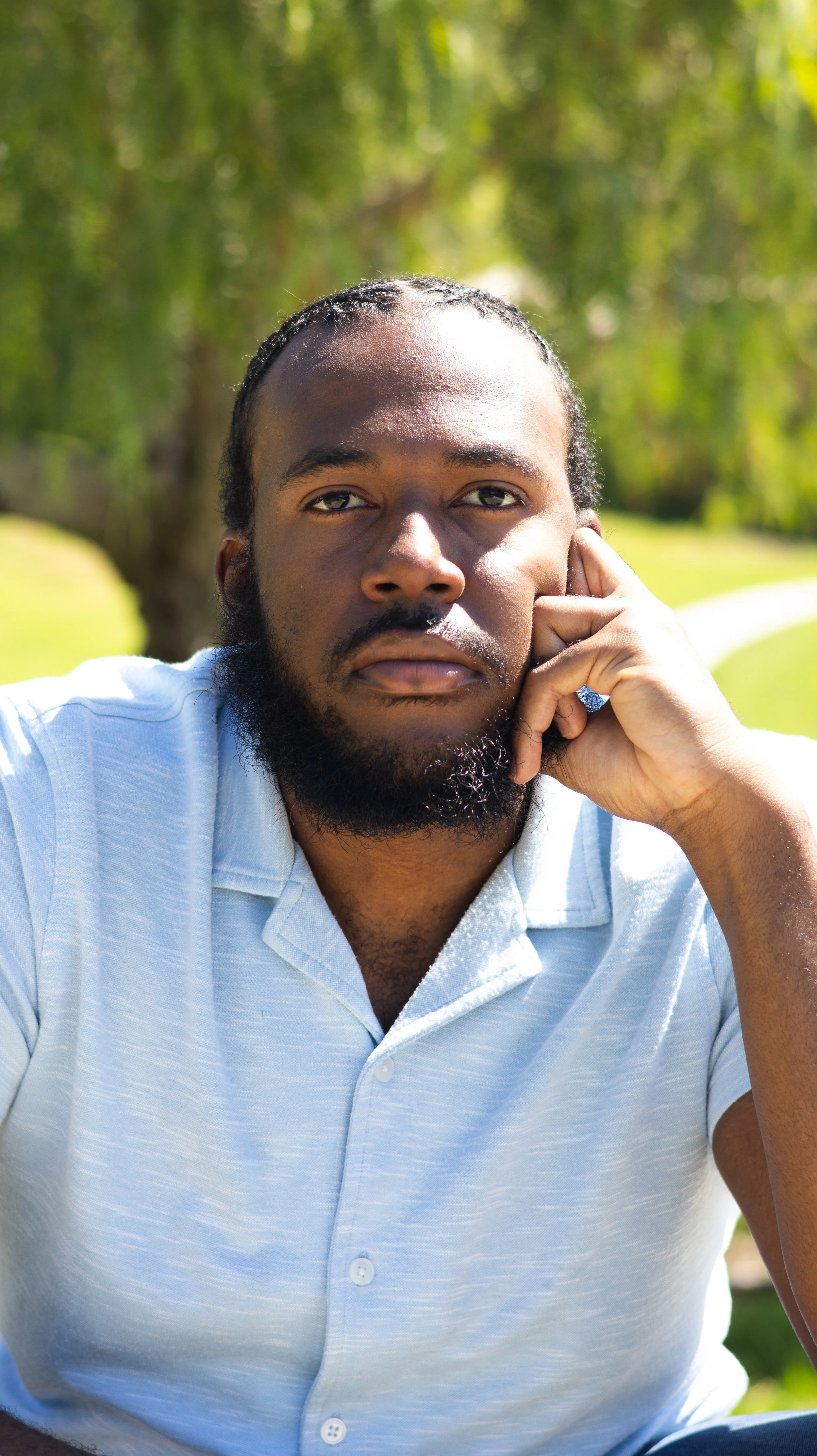 A man with a beard and long hair pulled back, wearing a light blue polo shirt, sitting outdoors on a sunny day with green trees and grass in the background.