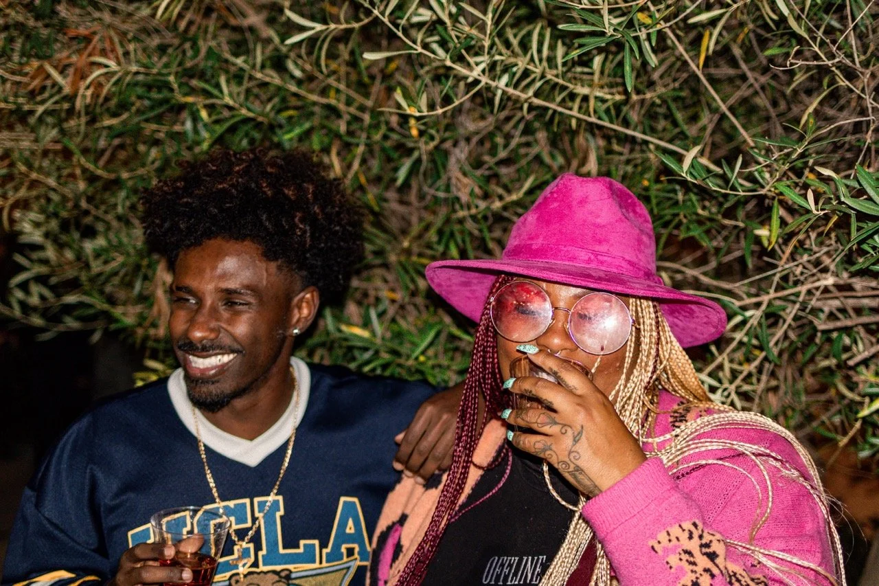 Two people smiling and laughing outdoors at night, one wearing a pink hat and glasses, and the other holding a glass of drink.
