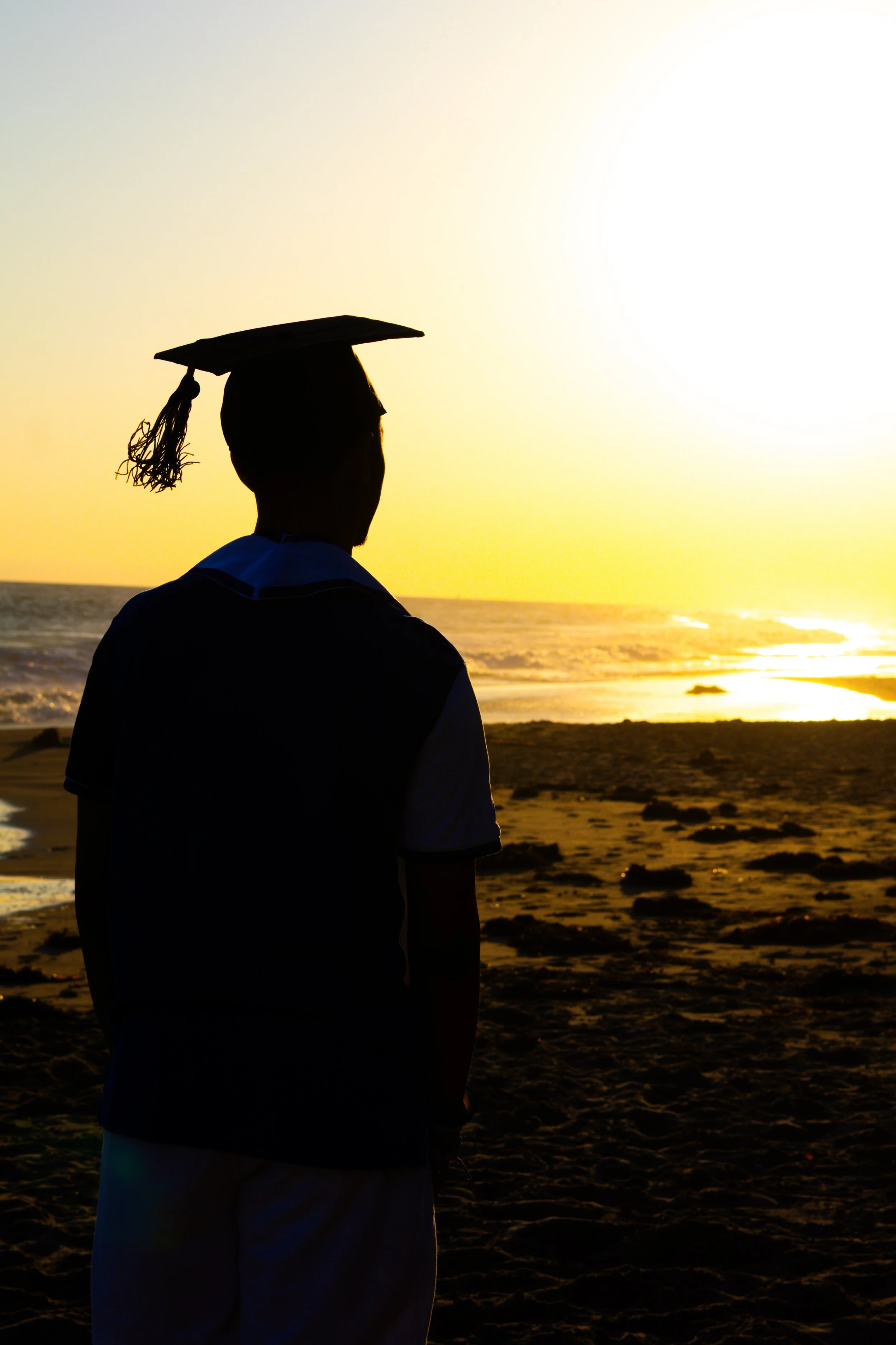 Silhouette of a person in a graduation cap and gown standing on the beach during sunset.
