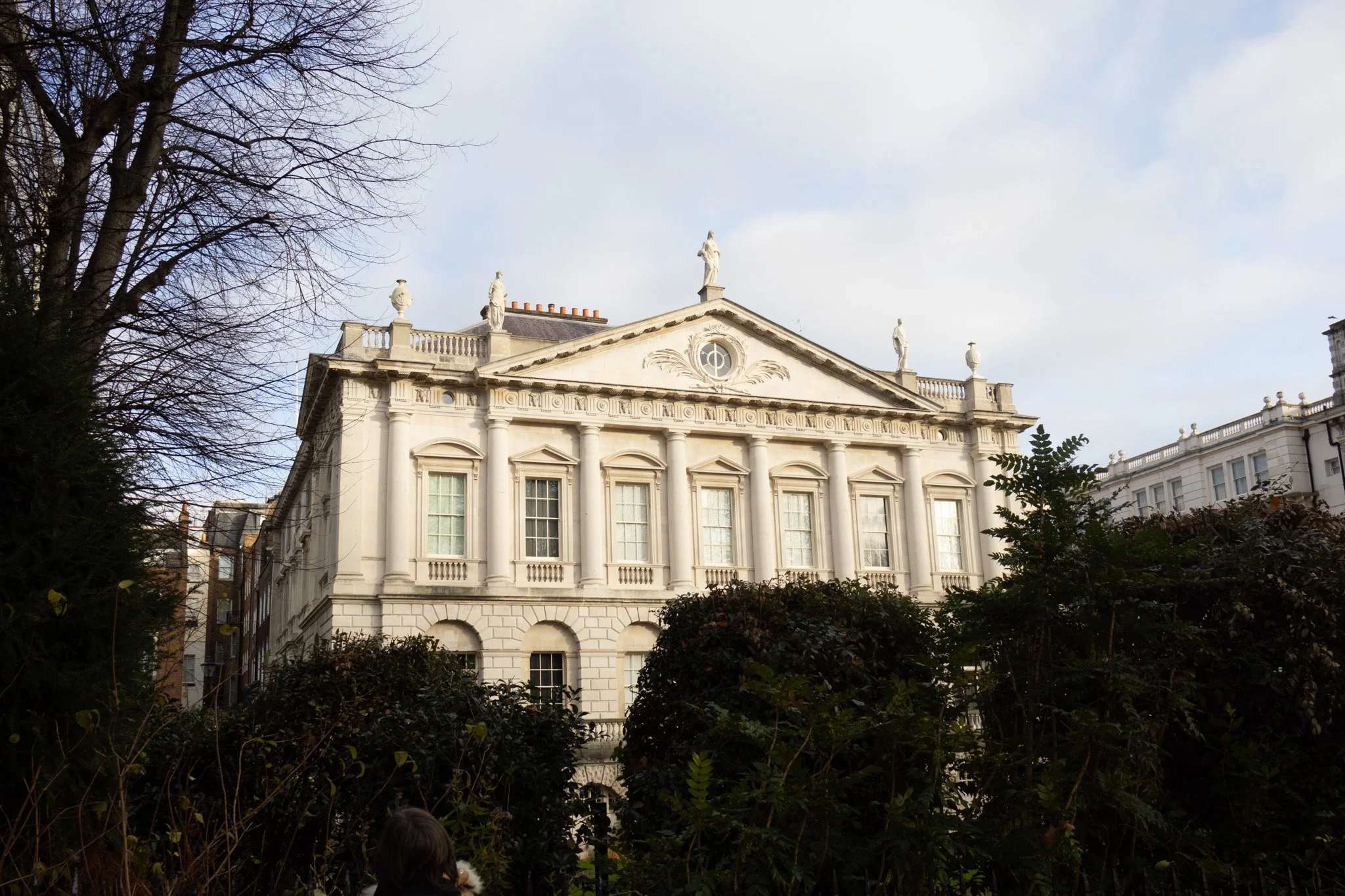 A white building with classical architecture, including columns and statues on the roof, surrounded by trees and bushes, under a cloudy sky.