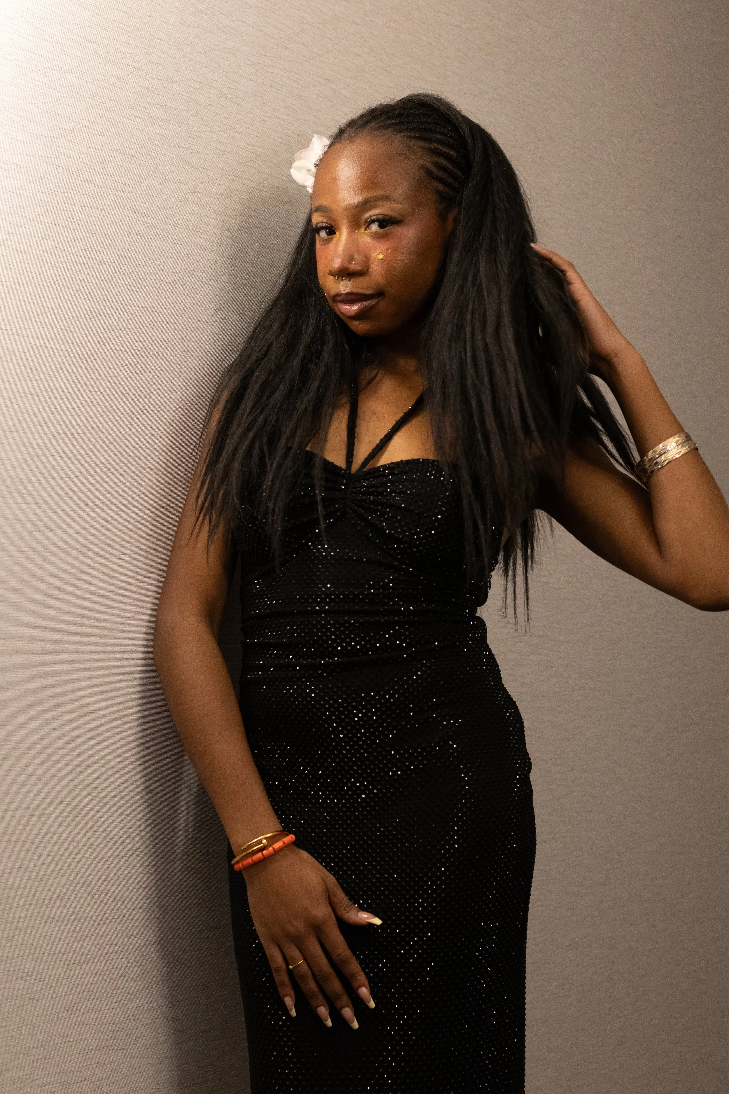 A young woman with long black hair and a flower hair accessory, wearing a black, sparkly strapless dress, standing against a gray wall.