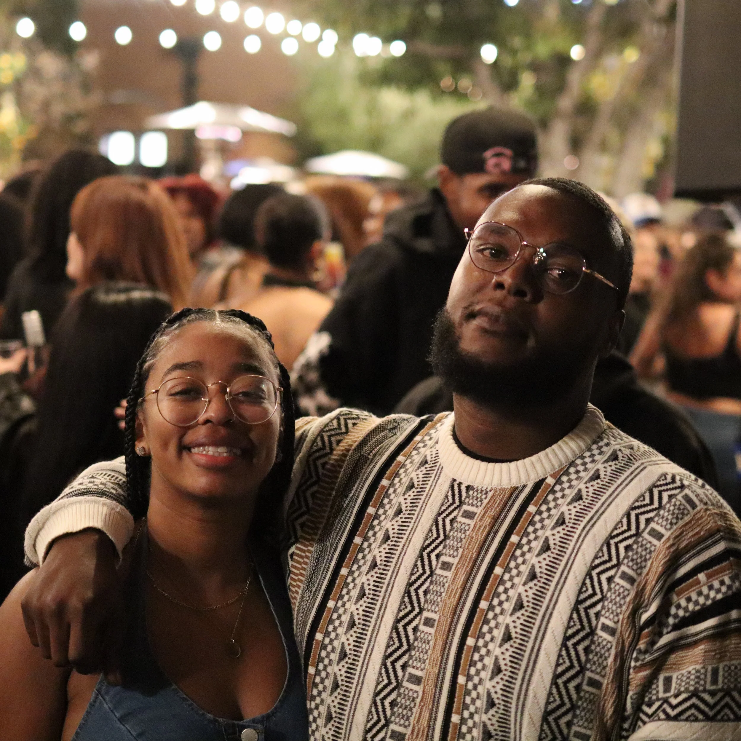 A smiling woman and man with glasses posing together at a crowded outdoor event at night.
