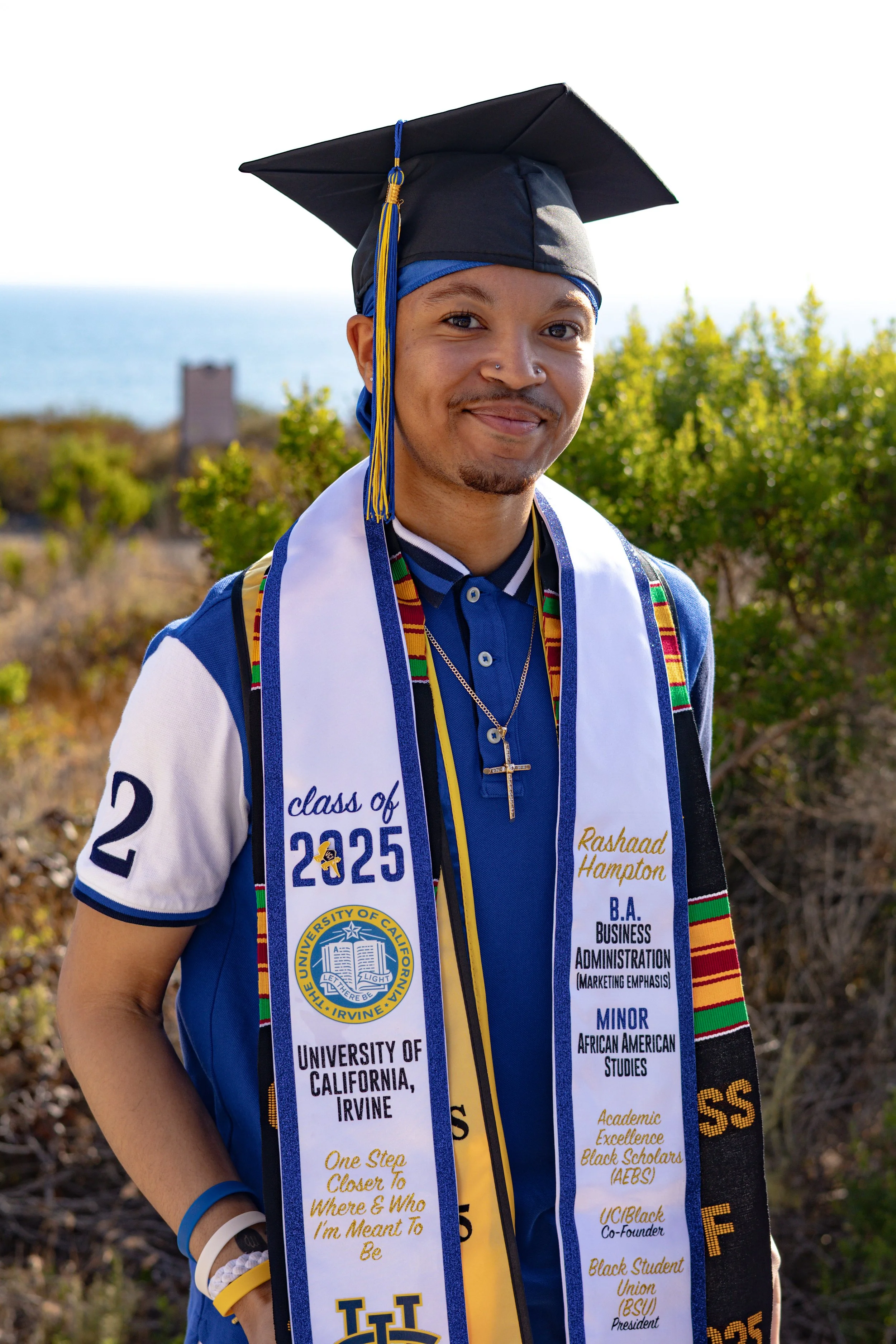 Young man celebrating graduation outdoors, wearing a cap, gown, and graduation stole from University of California, Irvine, with a background of greenery and a distant body of water.