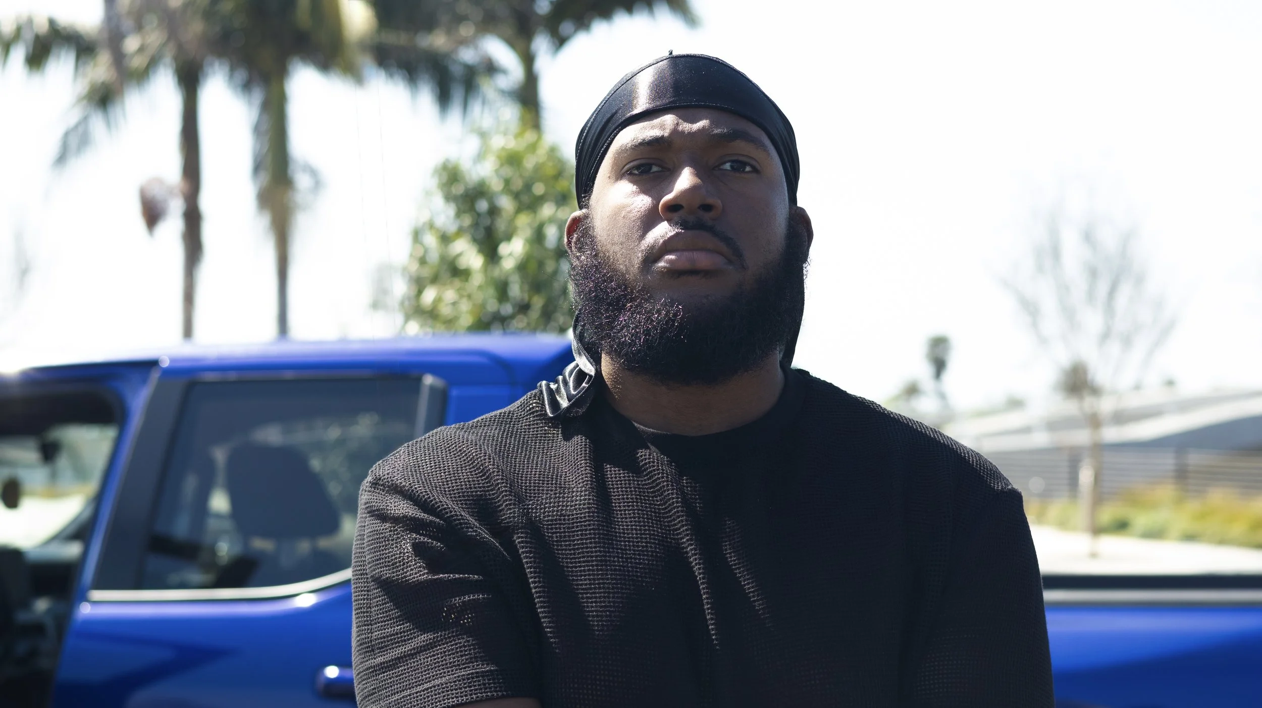 A man with a beard and black headwrap standing outdoors in front of a blue vehicle, with palm trees and a cloudy sky in the background.