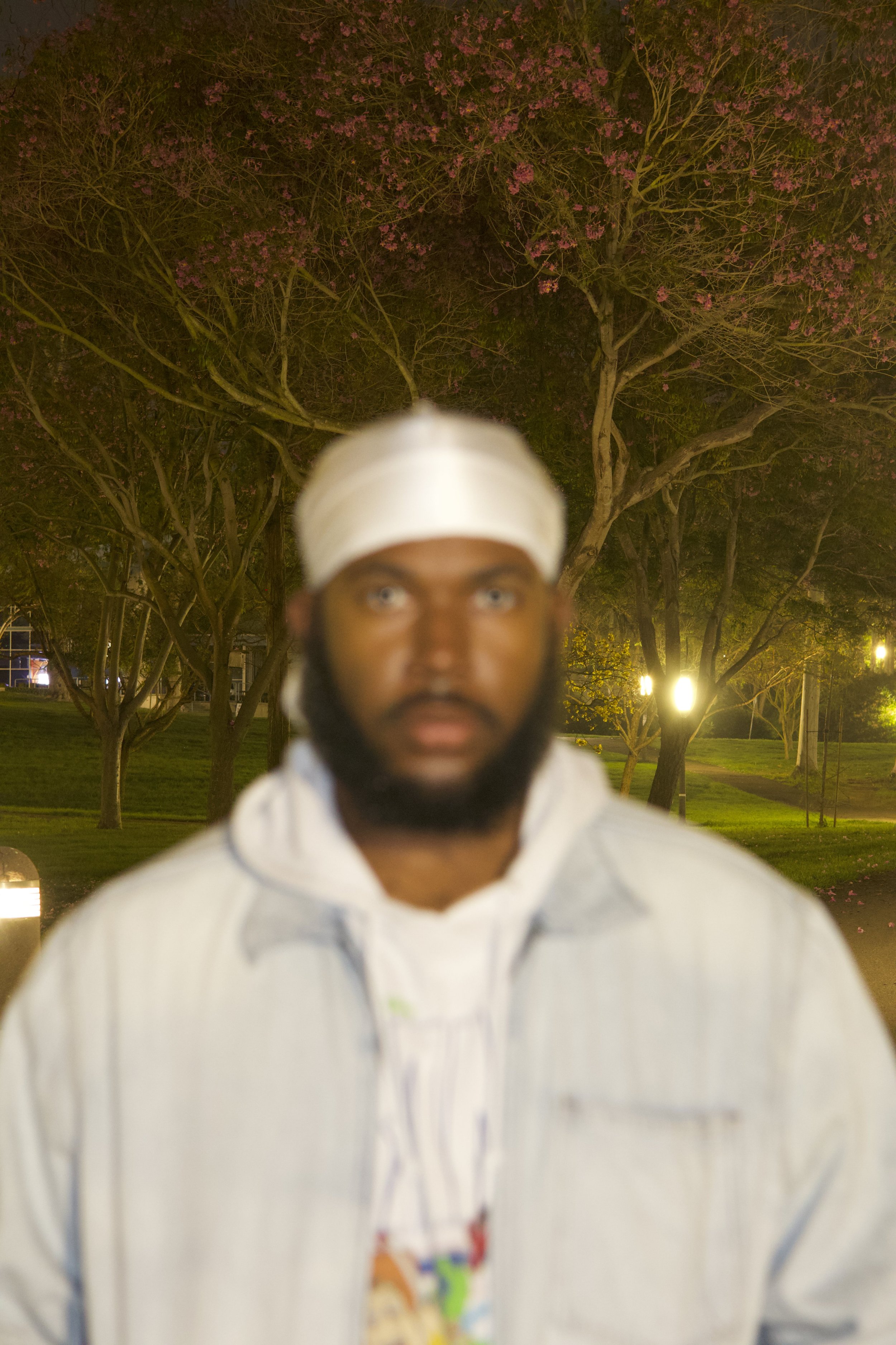 A man with a beard and wearing a white head wrap and white jacket standing outdoors at night with trees and streetlights in the background.