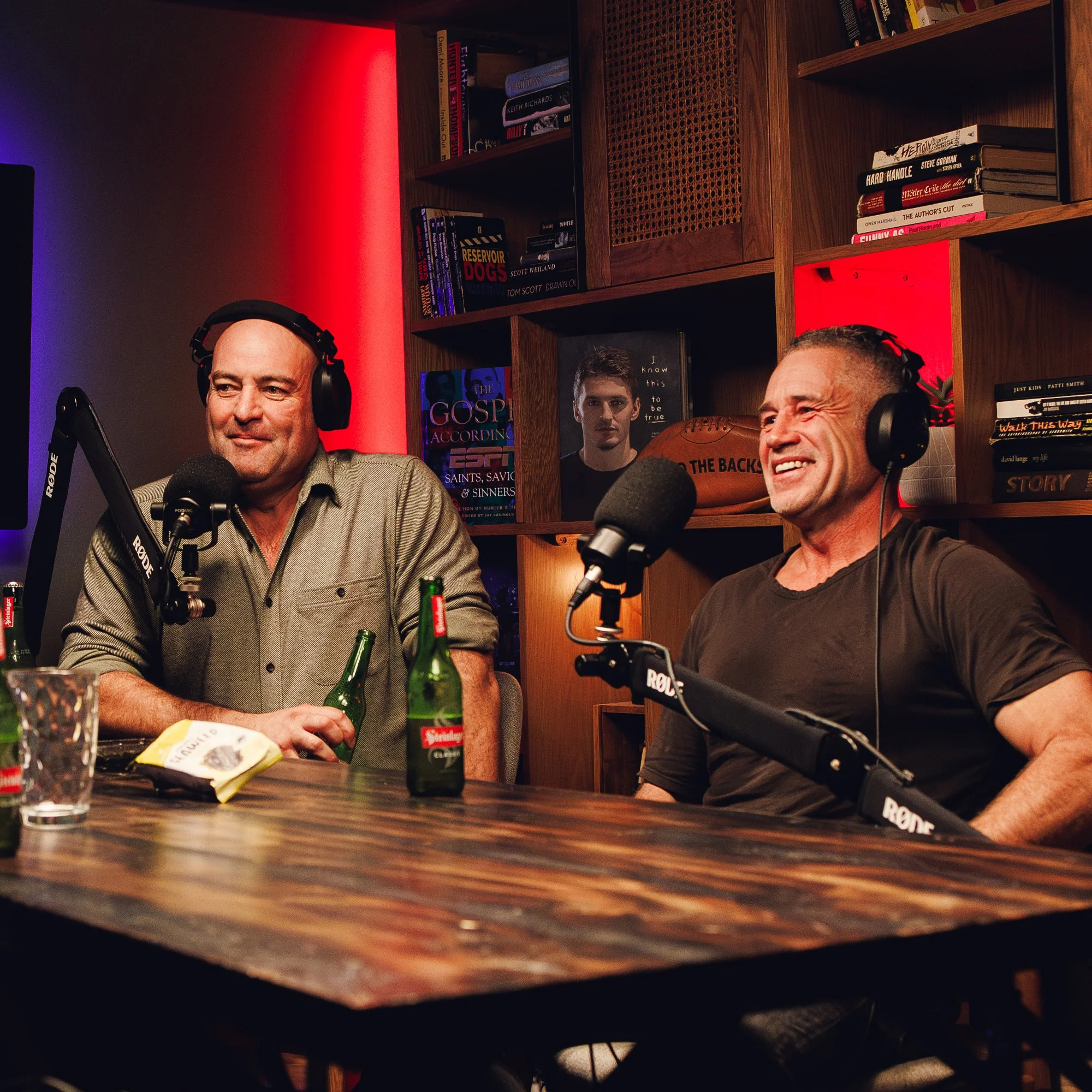 Two men wearing headphones sit at a podcast recording studio with bookshelves in the background and microphones in front of them.