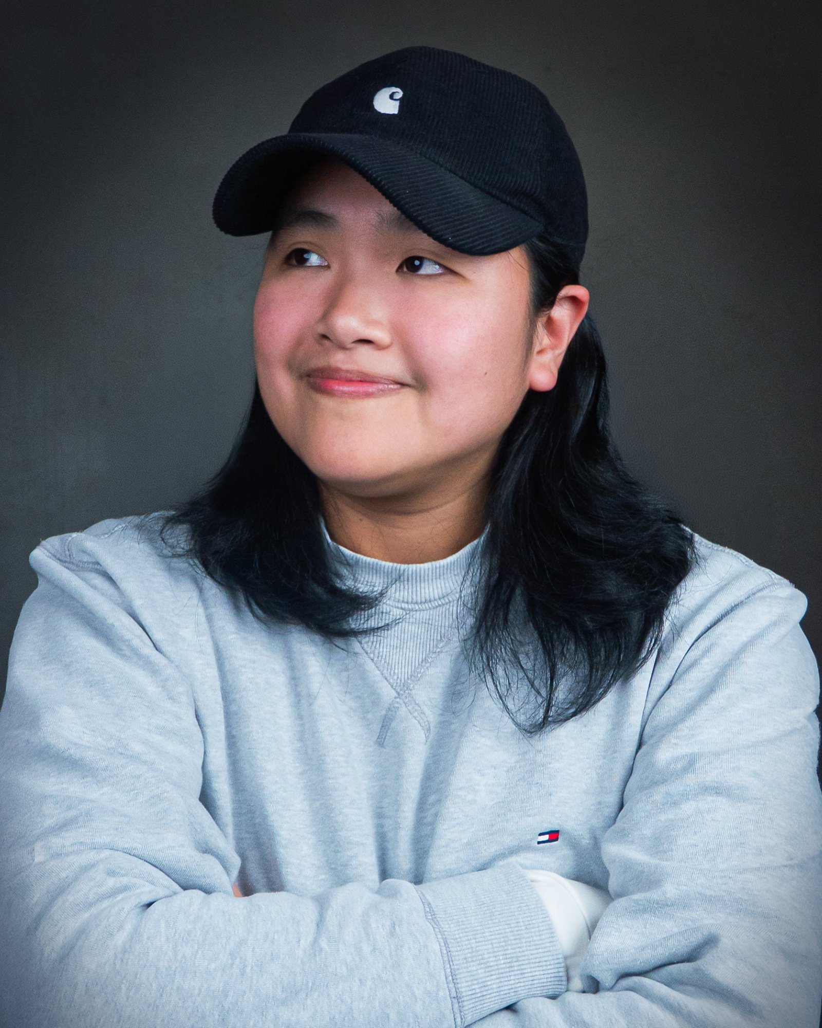 A person wearing a black baseball cap with a white logo, a light gray sweatshirt, and has long dark hair. The person is squinting and has their arms crossed in front of them, against a dark background.