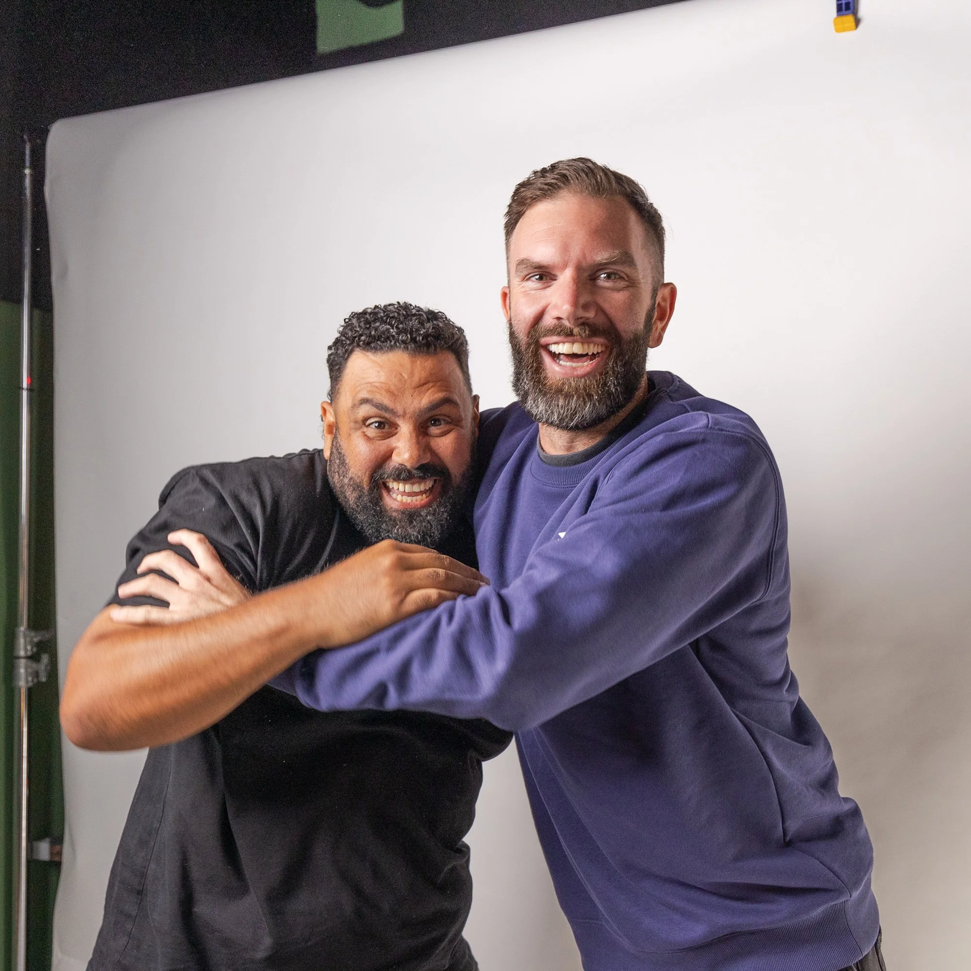 Two men smiling and hugging each other for a photo in a studio. The man on the left has short curly black hair, a beard, and is wearing a black shirt. The man on the right has short brown hair, a beard, and is wearing a dark purple shirt.