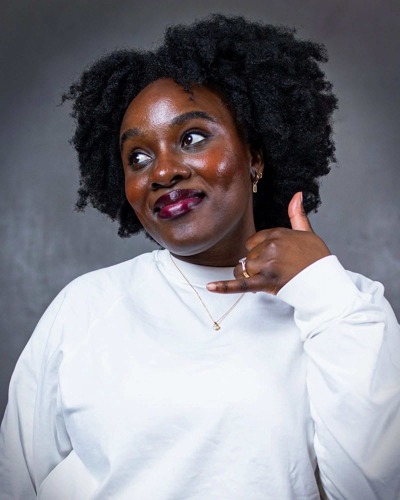 Close-up of a Black woman with natural curly hair, wearing a white shirt, gold earrings, and a gold necklace, posing with a thoughtful expression against a gray background.