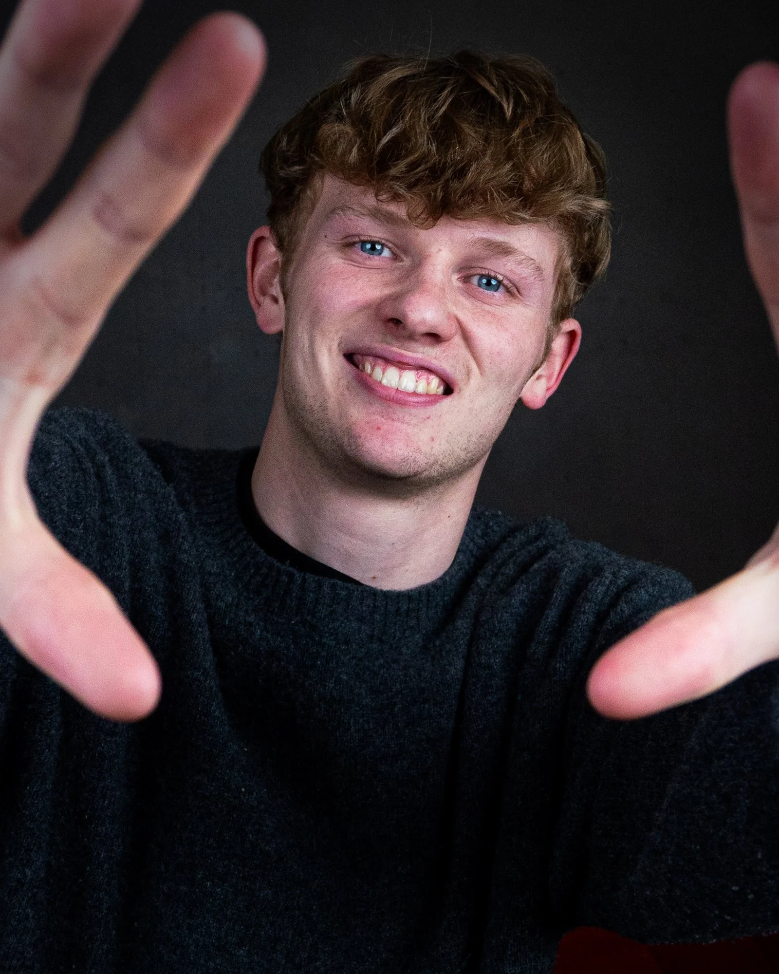 A young man with curly hair and blue eyes making a frame with his hands, smiling, against a dark background.