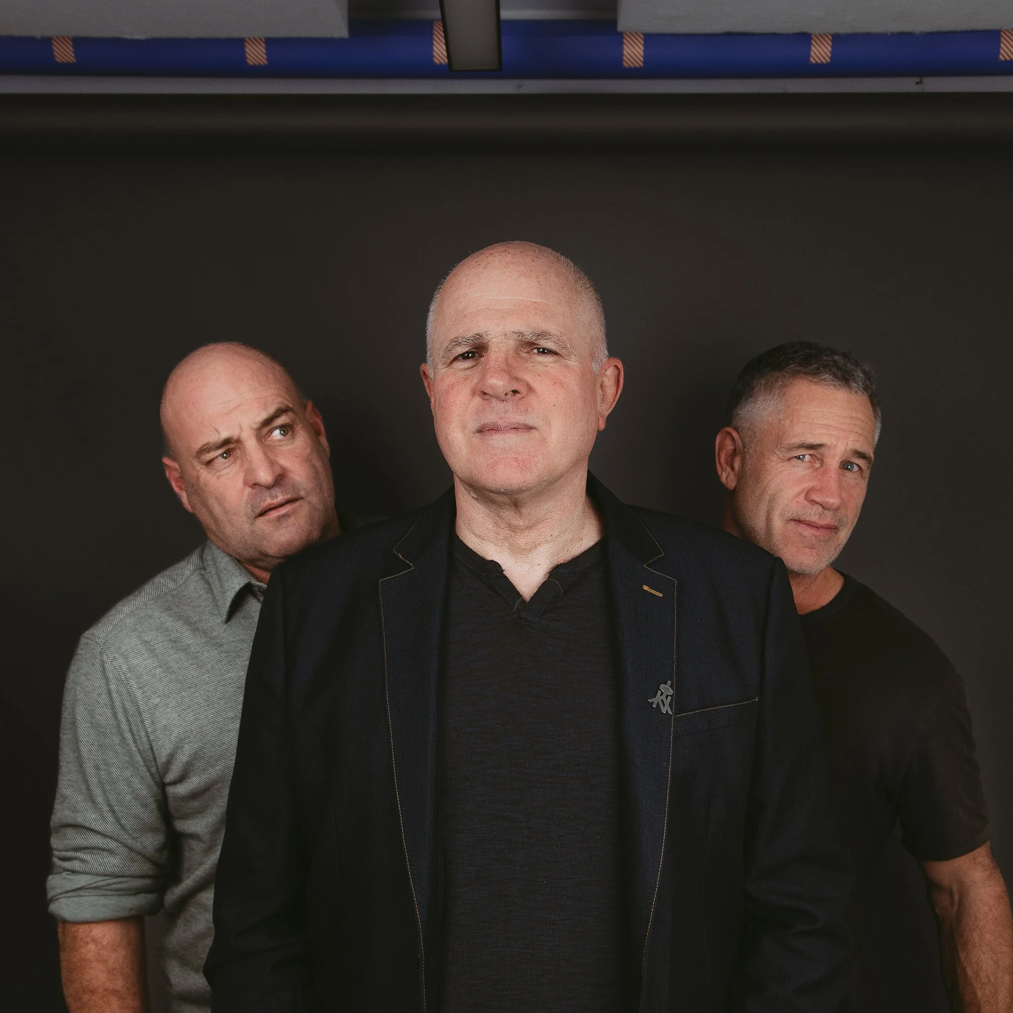 Close-up photo of five men standing together, with only their heads and shoulders visible, against a dark background. The men have different facial expressions and hairstyles.