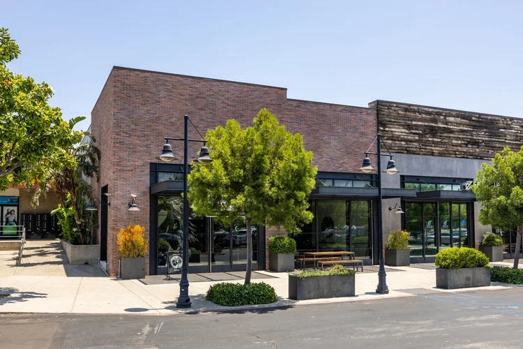 A modern commercial building with large glass windows, surrounded by trees and outdoor planters, next to a parking lot.
