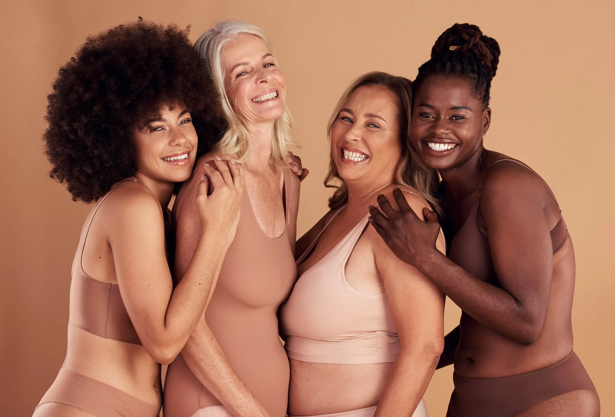 Four women of diverse ages and ethnic backgrounds smiling and hugging against a beige background, wearing nude-colored undergarments.