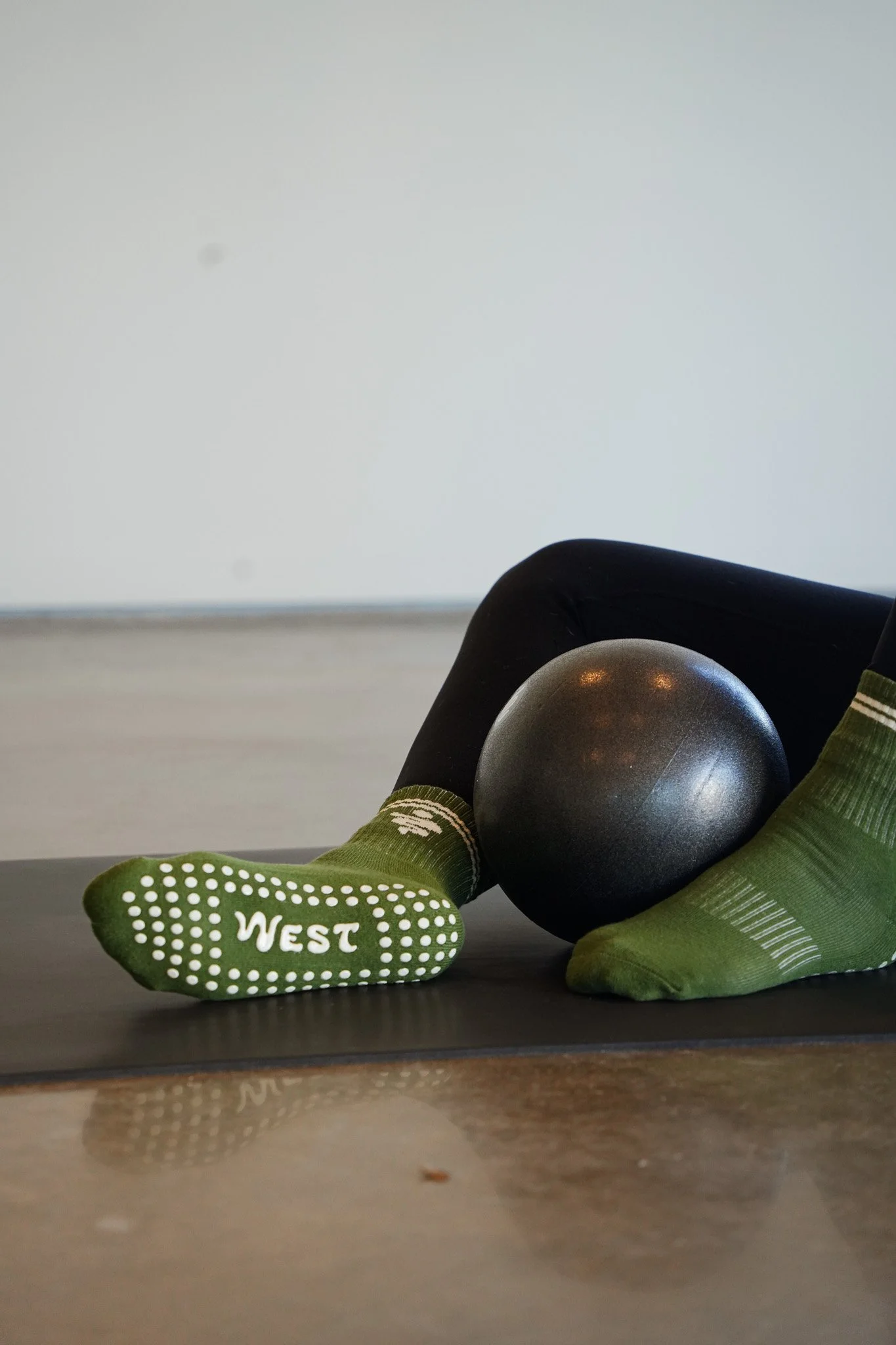 A small black kettlebell resting on its side on a gray surface. A green sock with white dots and the word 'WEST' printed on the sole is partially covering the kettlebell. The background is plain light gray.