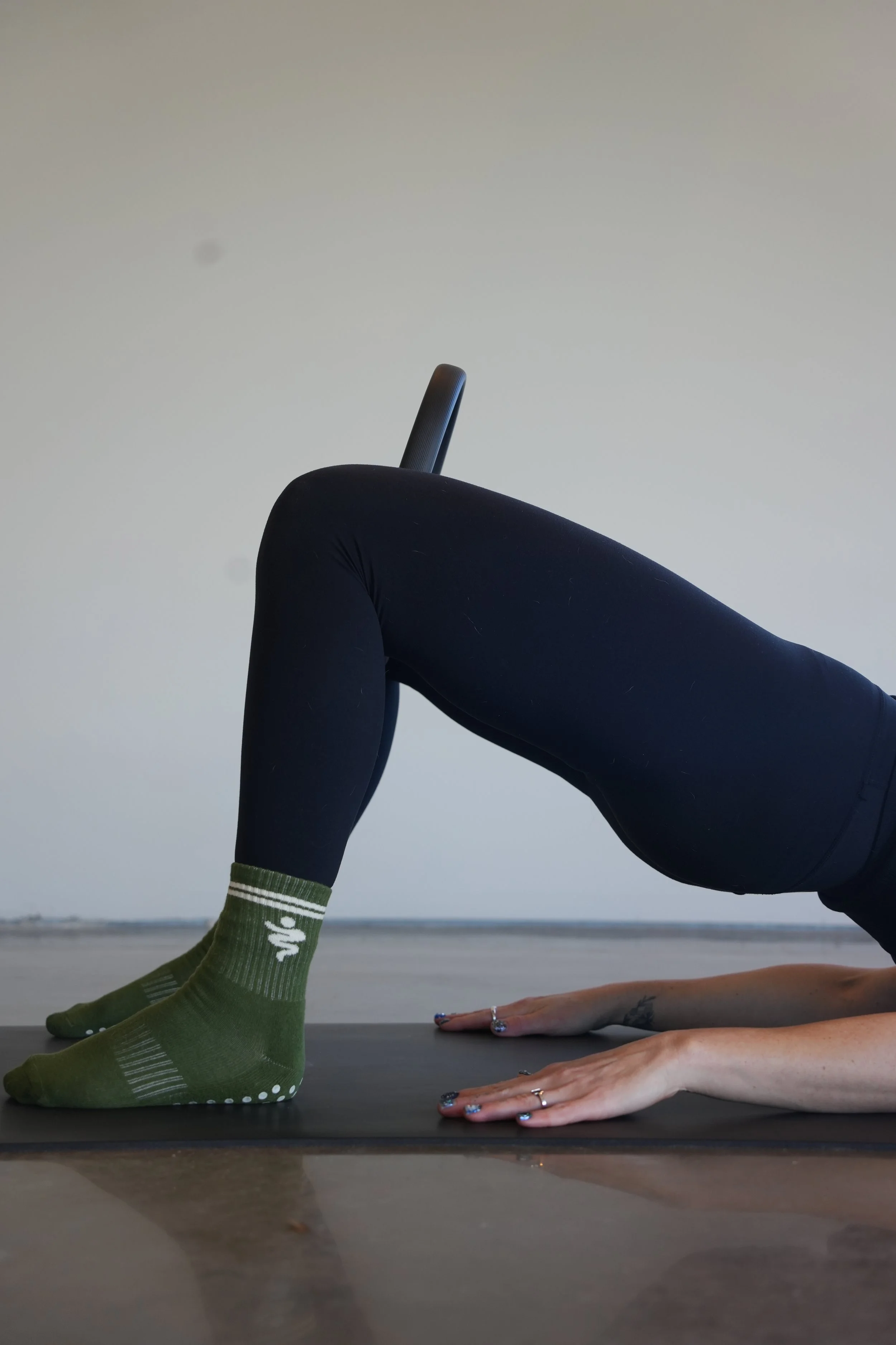 Person practicing yoga in an indoor studio, performing the downward dog pose with one leg raised, wearing black leggings, green socks, and rings on their hands, against a neutral background.