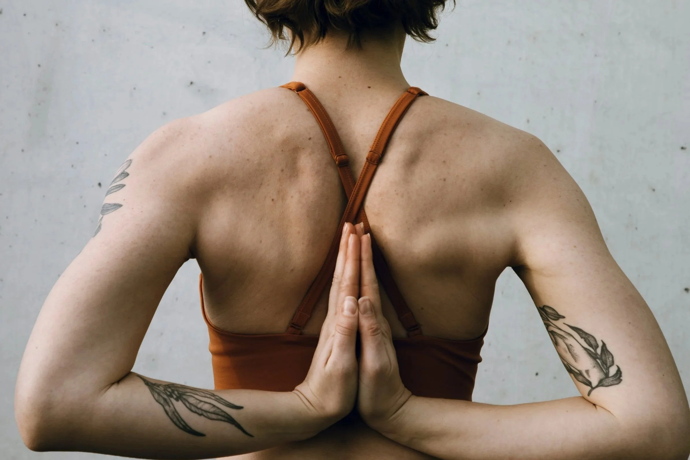 A woman practicing yoga outdoors, in a prayer pose with hands pressed together at her chest, wearing a brown sports bra, showing tattoos on her arms and shoulders, against a plain outdoor wall.
