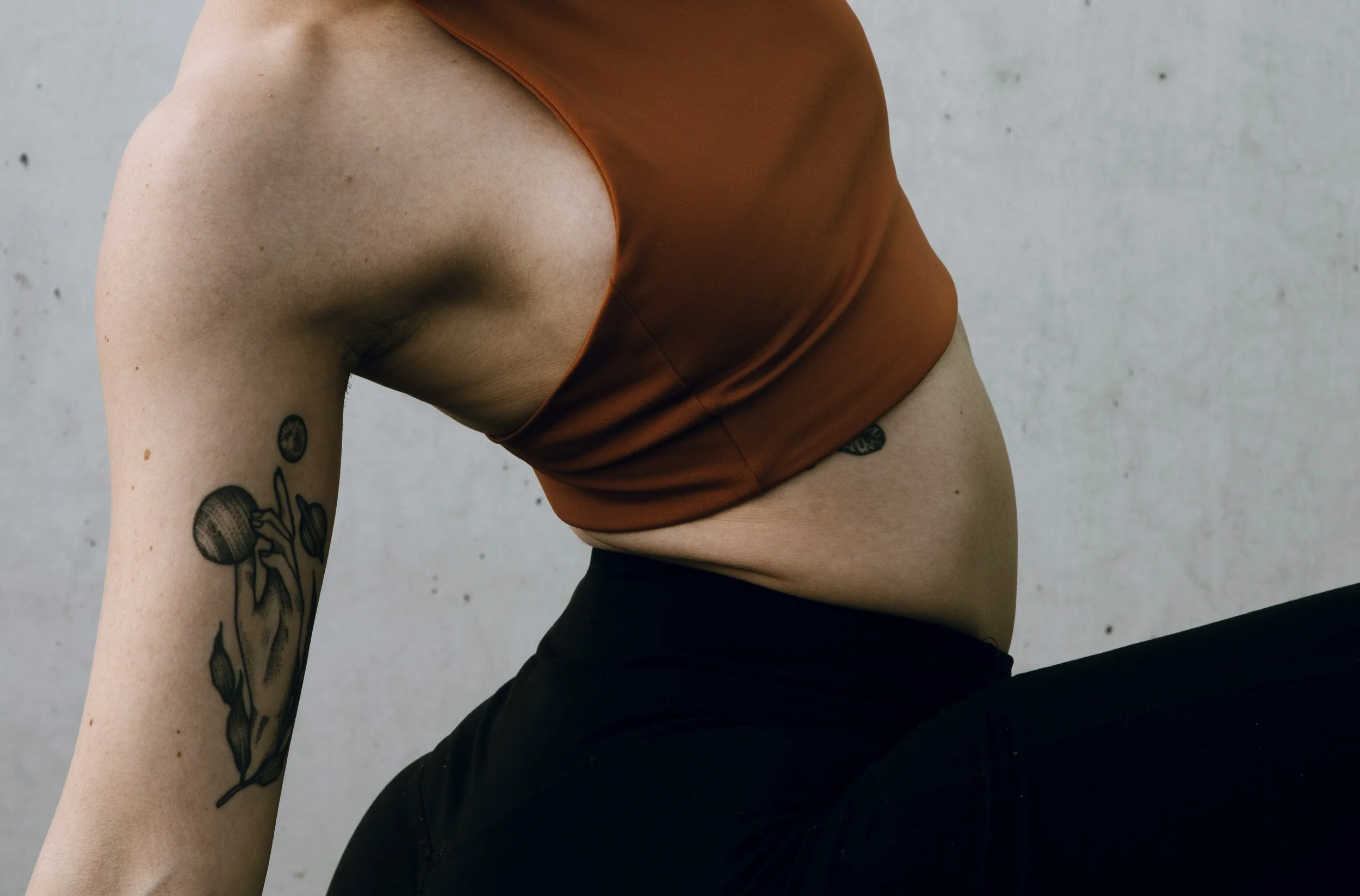 Close-up of a person wearing a rust-colored tank top and black pants, with tattoos on their arm, squatting against a plain gray wall.