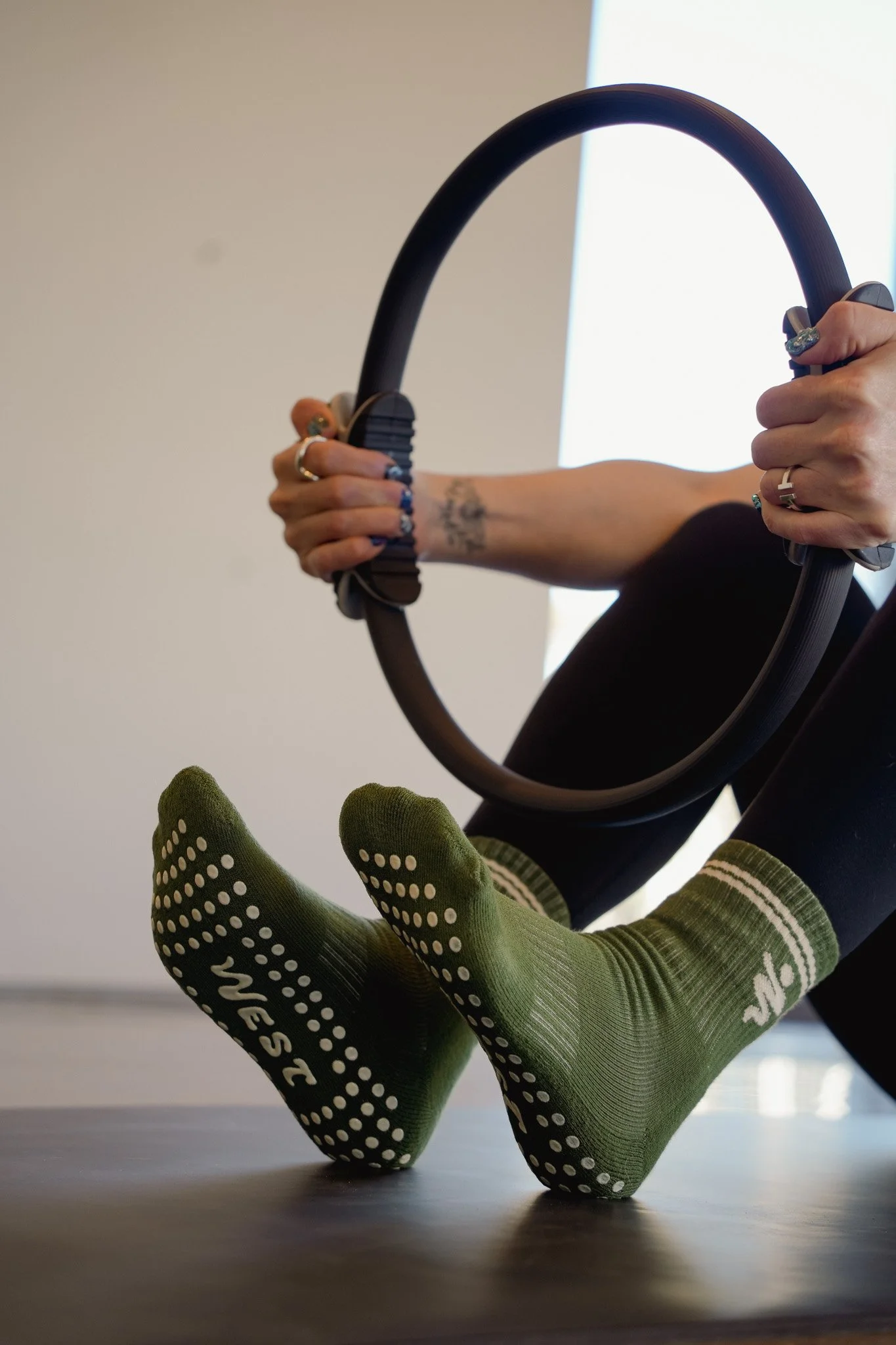 Two green socks with white dots and text on the bottom are worn by a person whose feet are propped up on a table. An individual is holding a large black fitness ring with both hands, with the background featuring a wall and window.