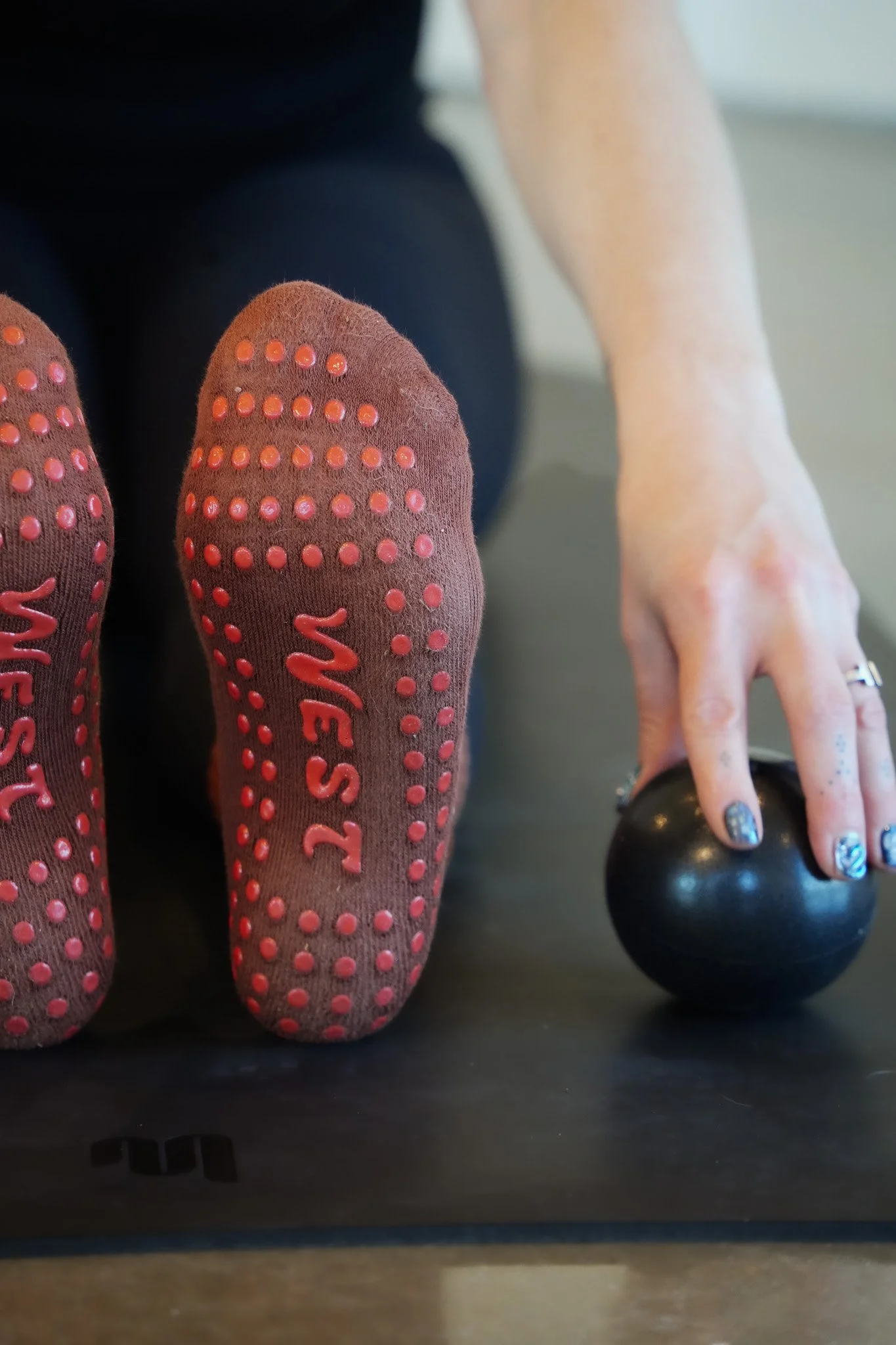 Close-up of person's feet in pink socks with small raised bumps and the word 'SASSY' on each sock. The person is holding a black kettlebell with their right hand, and part of their arm and hand are visible, with painted nails and a ring on the finger.