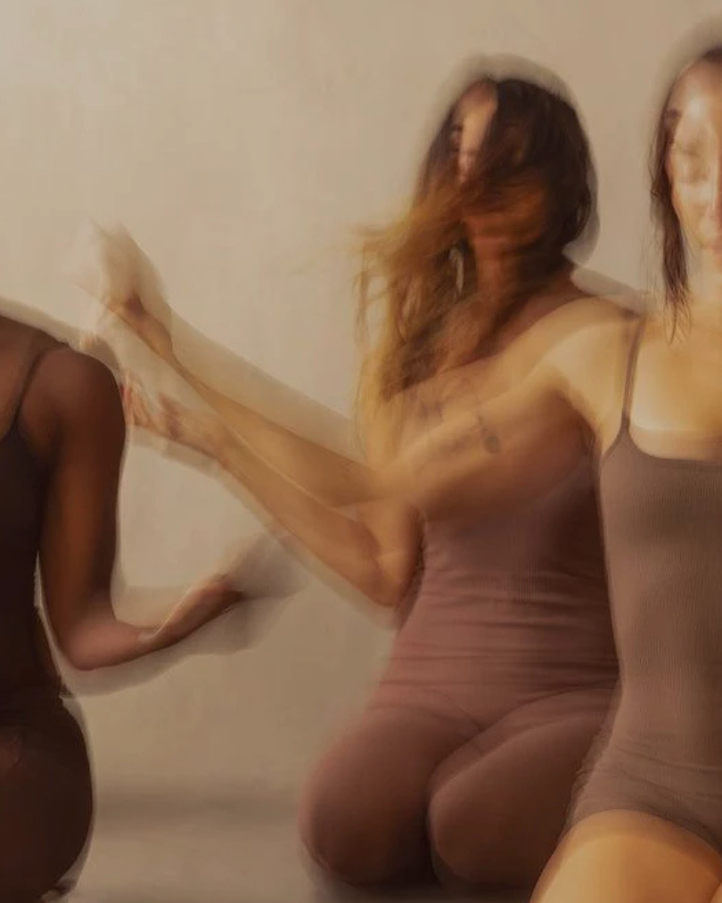Three women in sleeveless dresses pose together against a neutral background, with one woman having her face obscured by hair and motion blur.