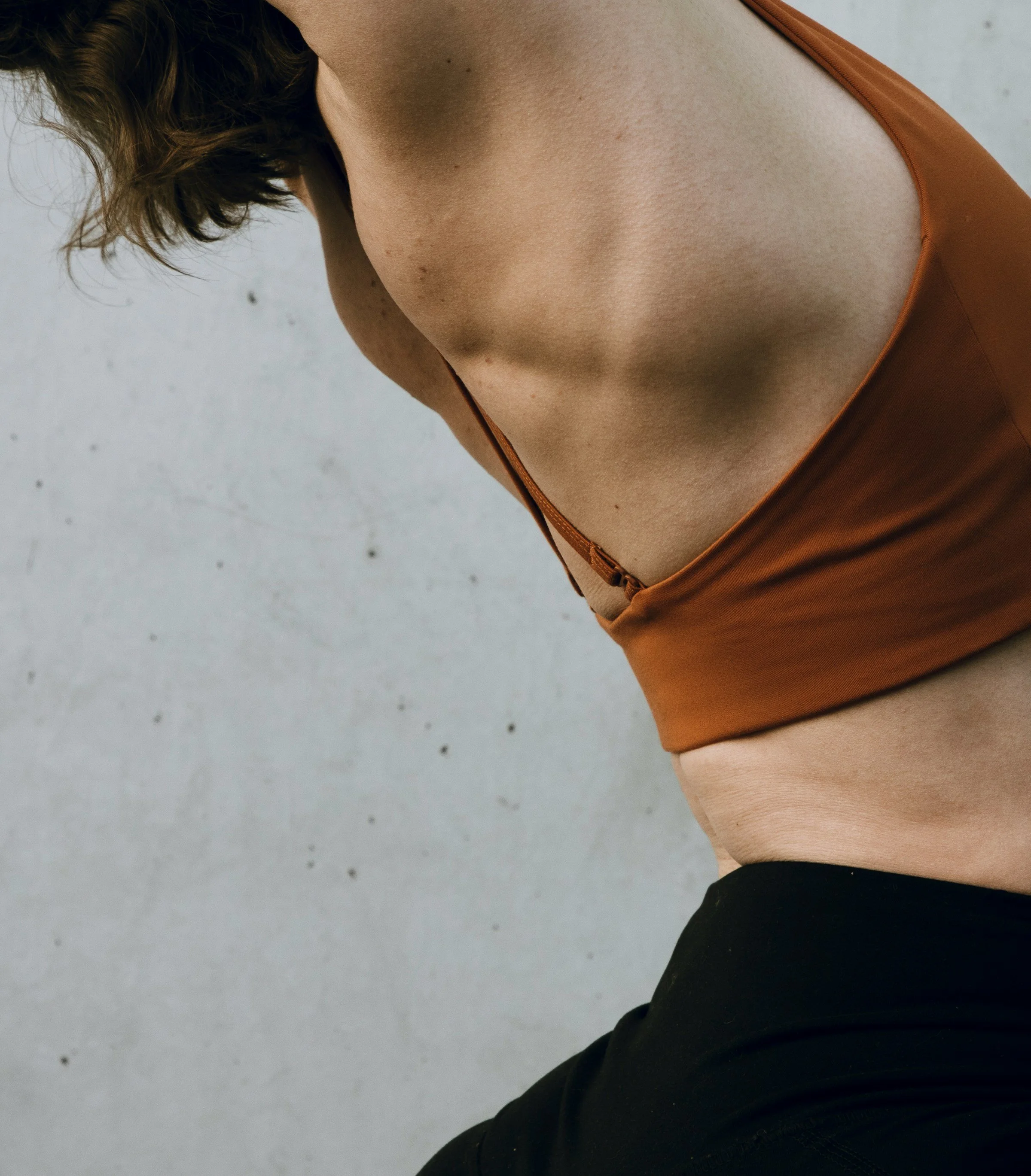 Close-up of a person wearing a brown tank top, with their shoulder and part of their arm visible, standing against a plain, light-colored background. Their head is turned away and slightly to the side.