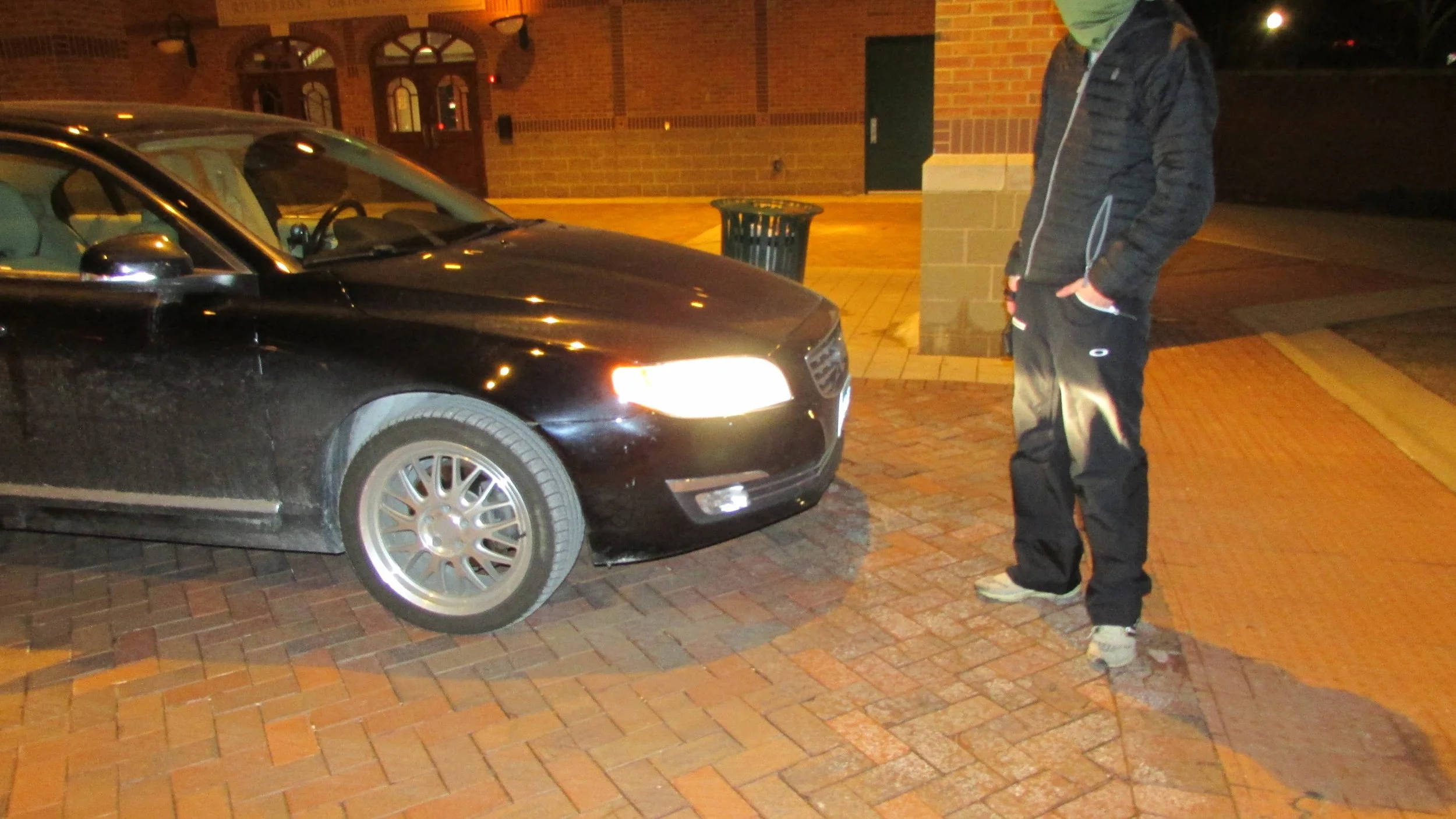 Ben stands on a brick sidewalk near a black car with headlights on, wearing a black jacket, dark pants, and gray shoes, with a green face mask.