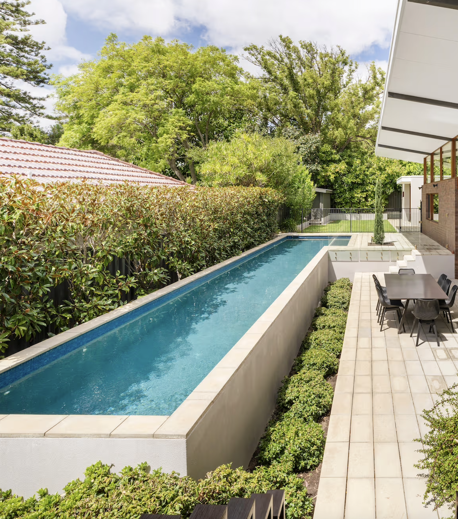A backyard with a rectangular swimming pool, surrounded by green shrubs and trees, with a patio area featuring outdoor chairs and a table, and a house with a balcony on the right.