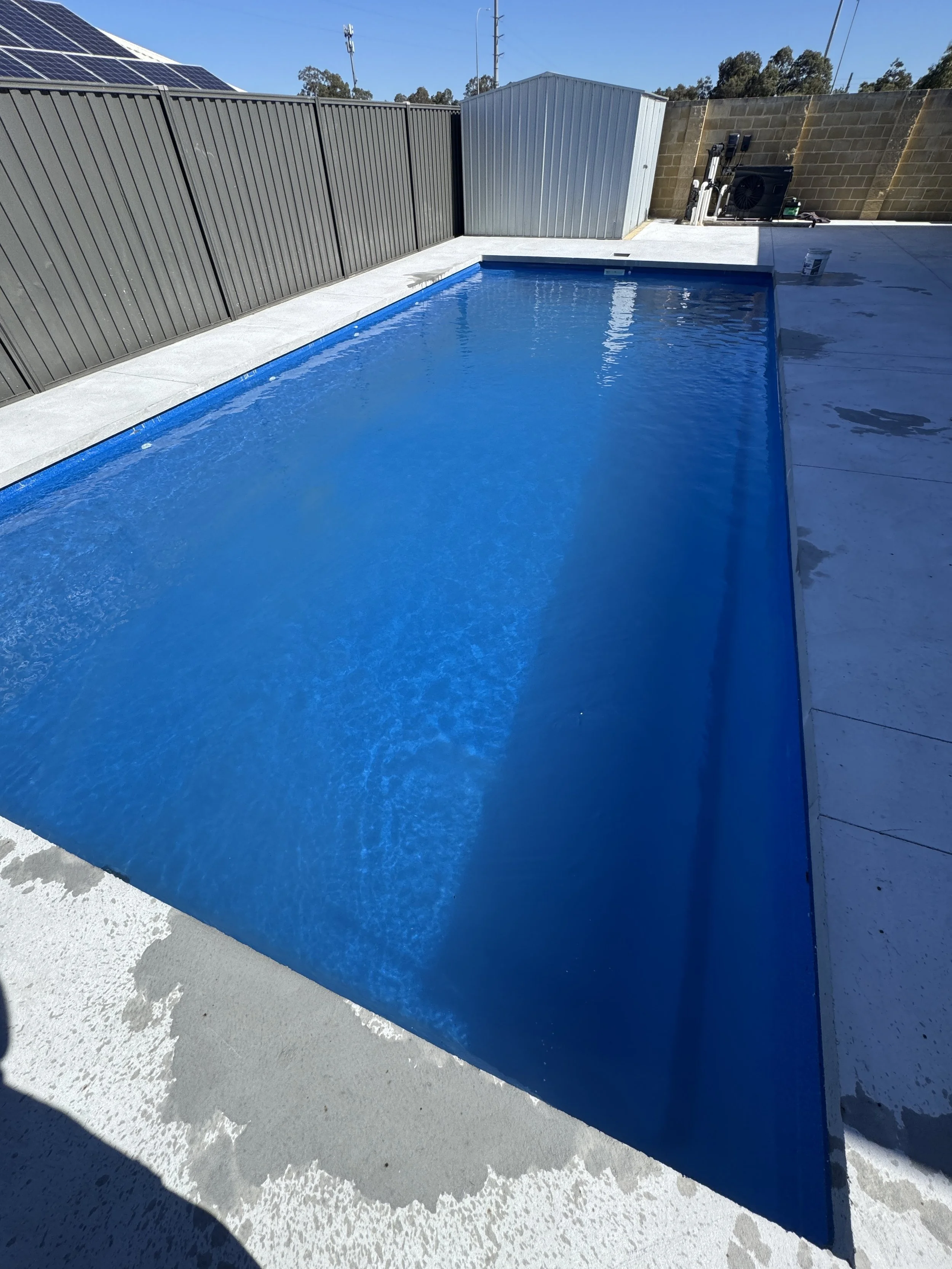 A backyard with a swimming pool, surrounded by a concrete deck and fenced with metal panels. There is a small shed or storage unit at the far end of the yard, with utility equipment and a brick wall in the background. The sky is blue and sunny.