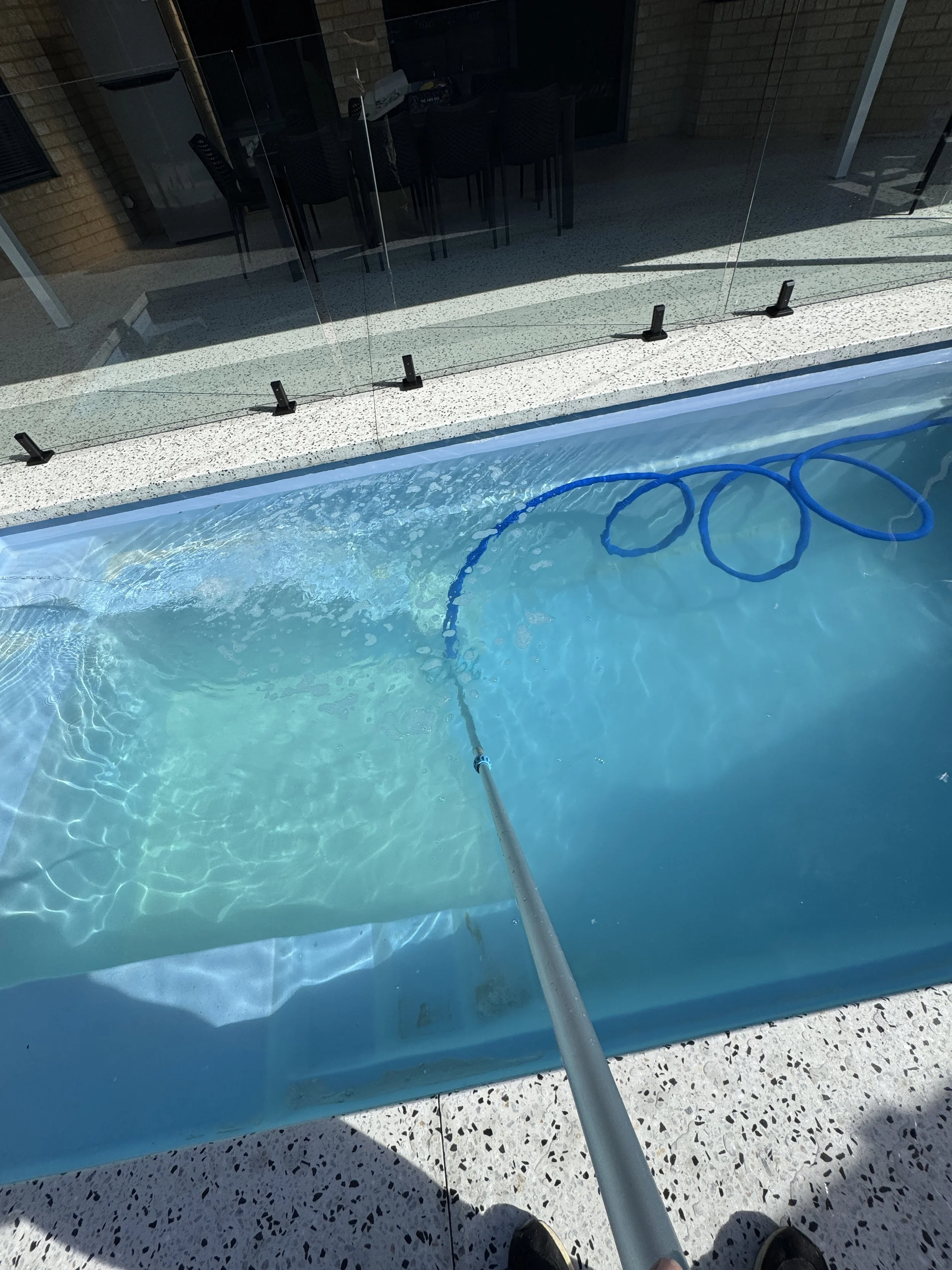 Person cleaning a glass pool fence with a pole and cleaning attachment, pool visible in the foreground, patio area with chairs and a table in the background.