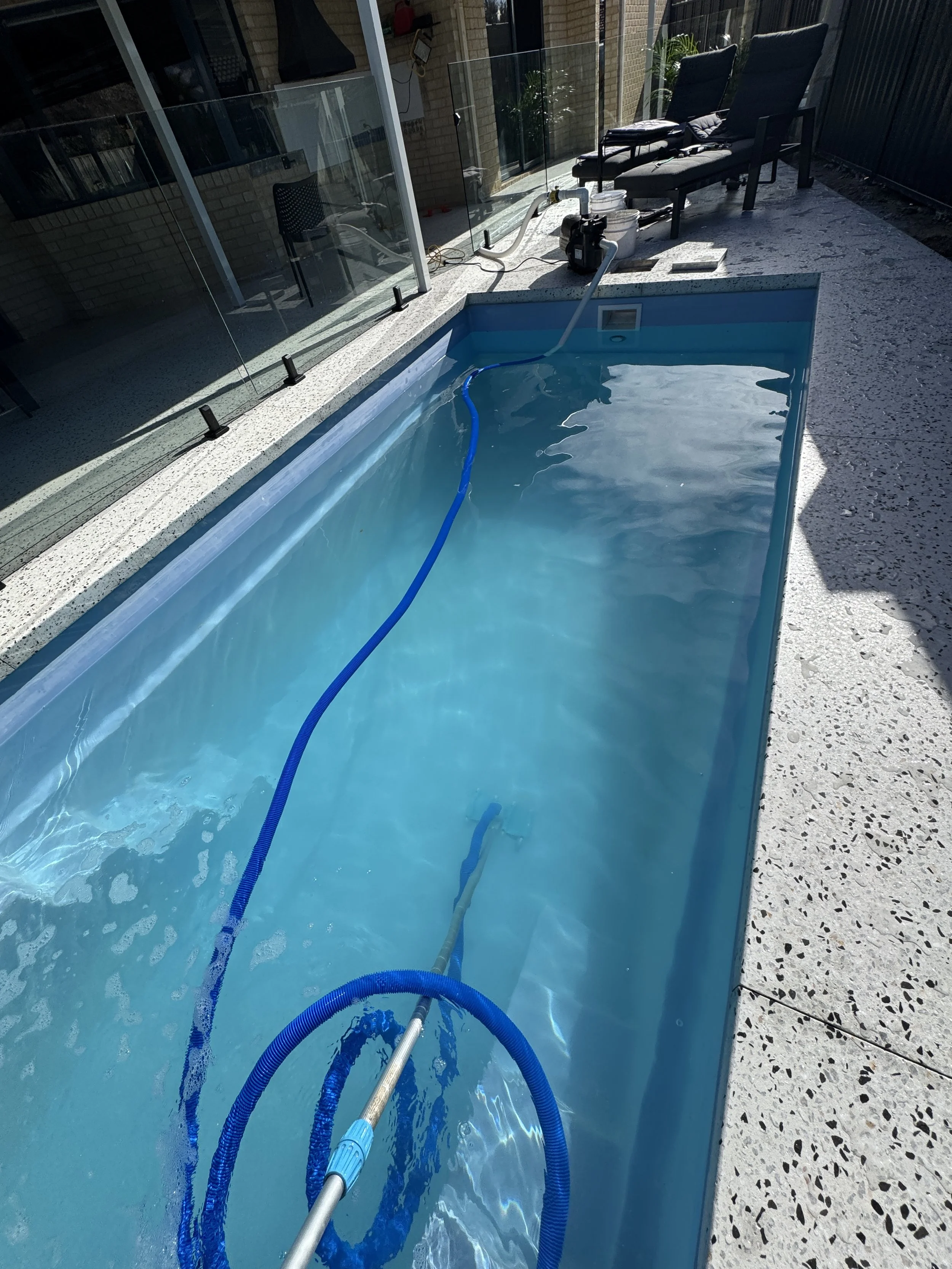 A swimming pool filled with blue water, with a blue hose and pole in the pool, near a pool cleaning or filtration device. There are two black chairs with cushions on a concrete patio next to the pool, and a glass safety fence separates the pool area from the house. The house has a brick exterior and large glass sliding doors.
