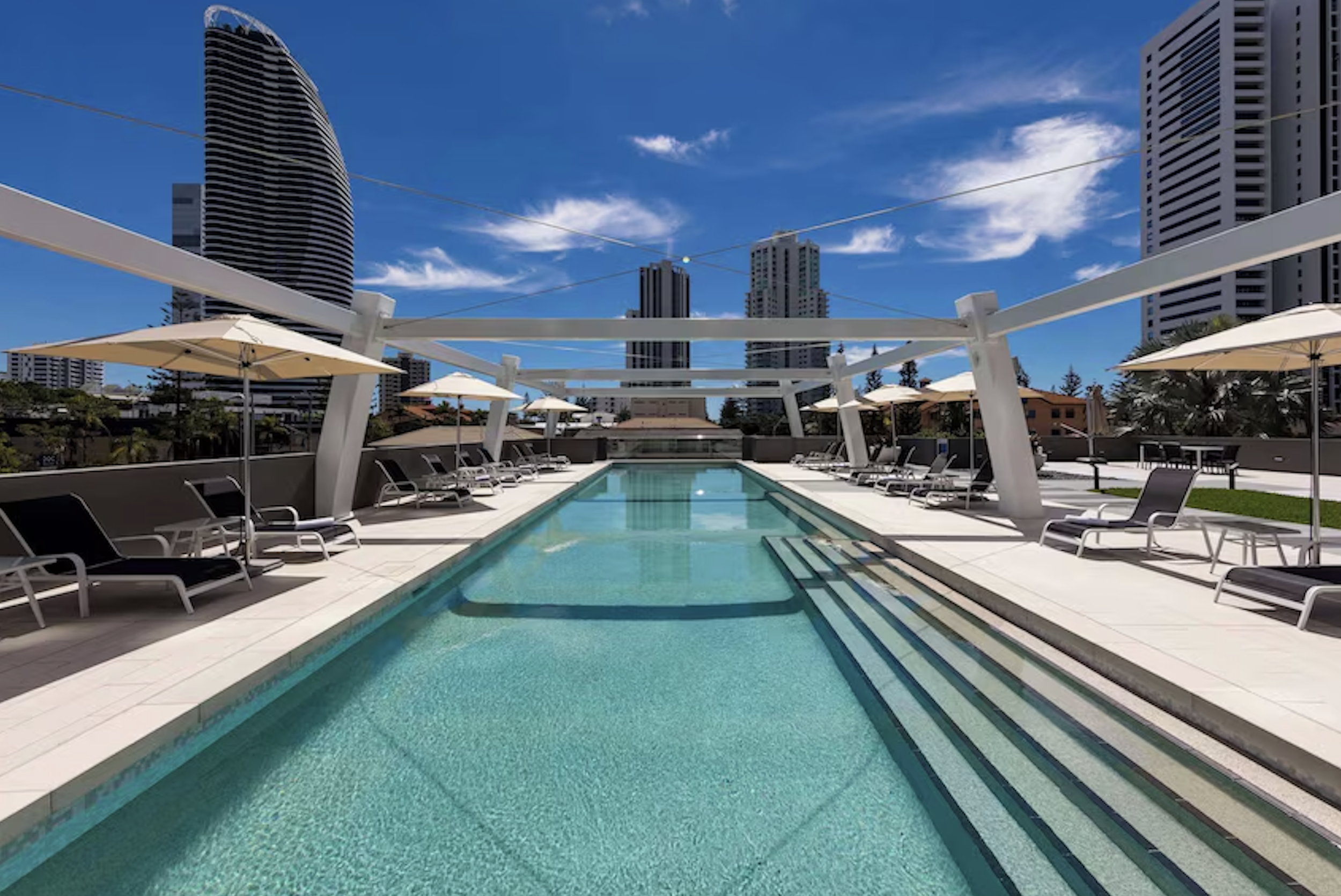 Urban rooftop pool area with lounge chairs, umbrellas, and a city skyline with high-rise buildings under a partly cloudy sky.