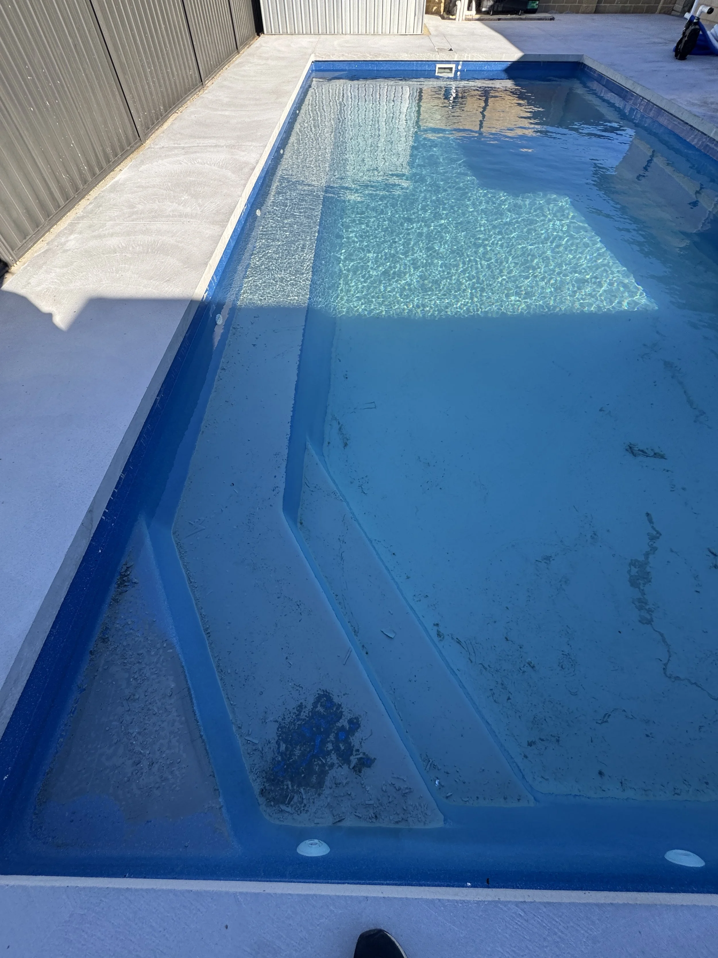 View of a backyard swimming pool with blue water, surrounded by a beige concrete deck, and partially shaded area with a dark fence and a building in the background.