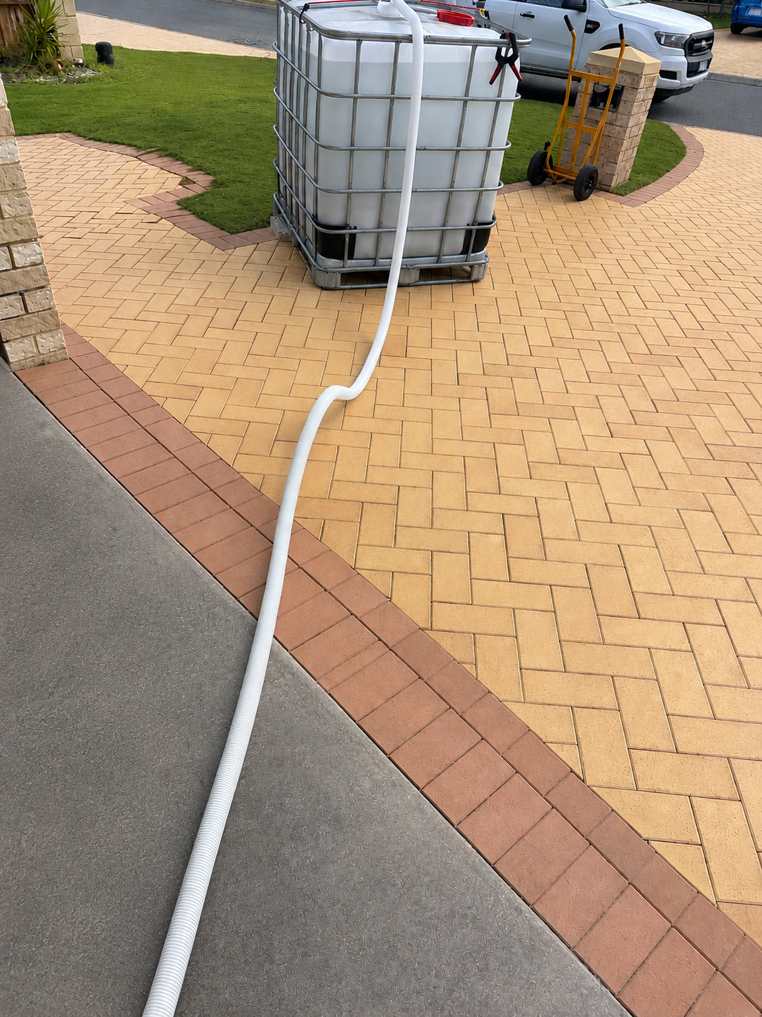 A large white water storage tank on a yellow brick patio, with a white hose connecting to it, near a grassy yard and parked vehicles in the background.